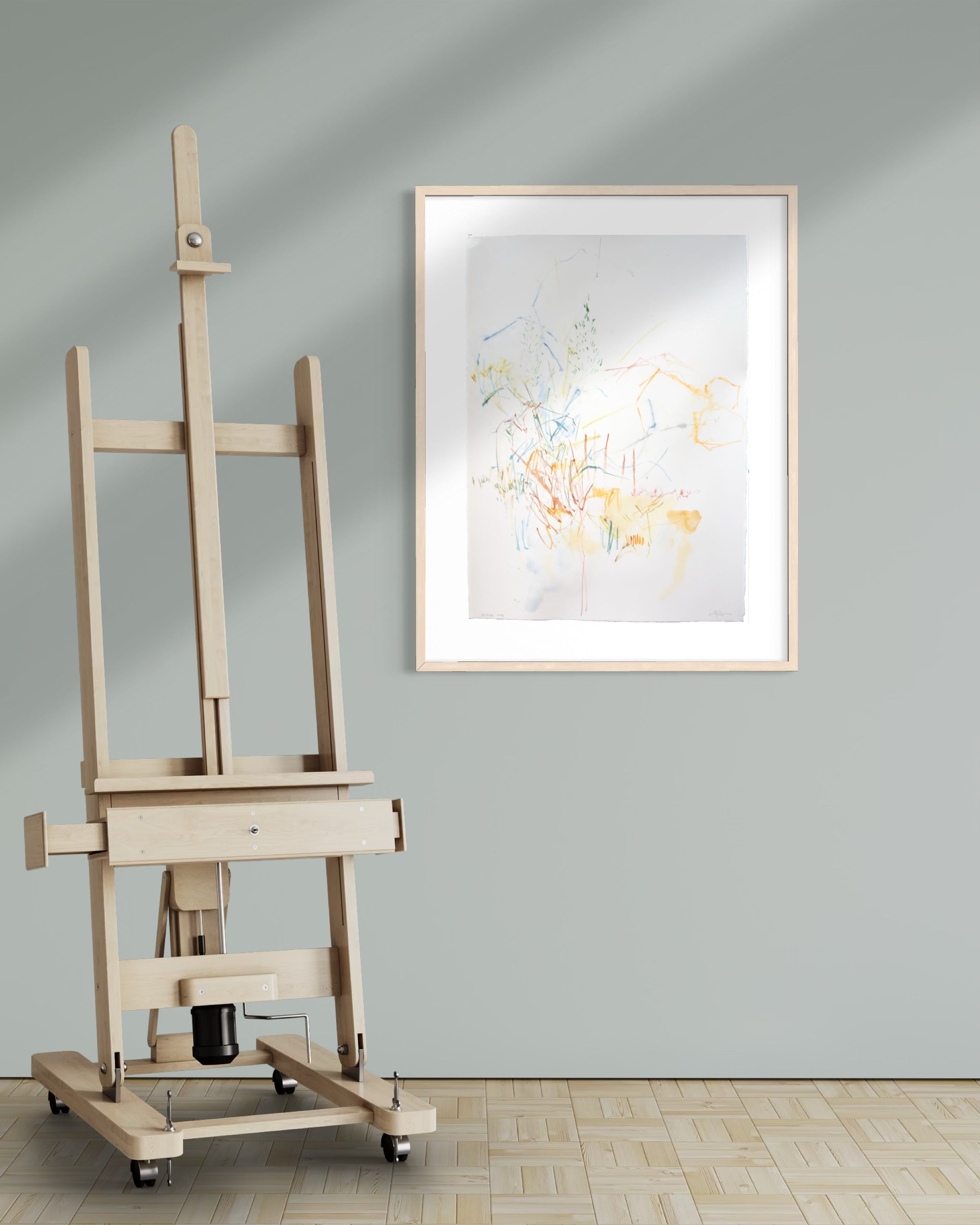 A light natural wood artist easel stands on a pale wood floor against a muted gray wall, next to a framed abstract art print with delicate yellow, blue, and green brushstrokes.