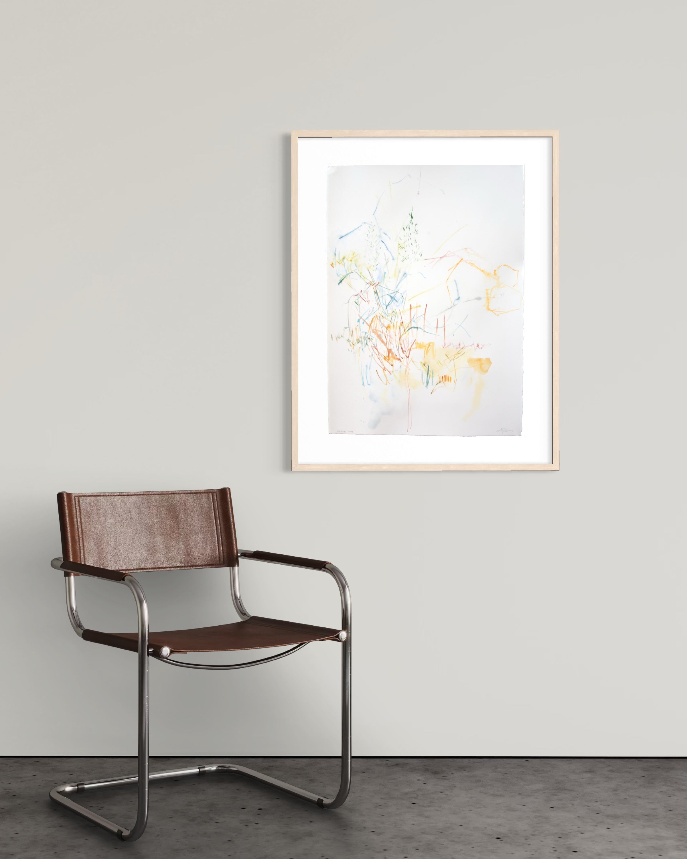 A framed abstract art print with delicate yellow, blue, and orange brushstrokes on a white background, styled on a light gray wall above a brown leather and chrome cantilever chair.