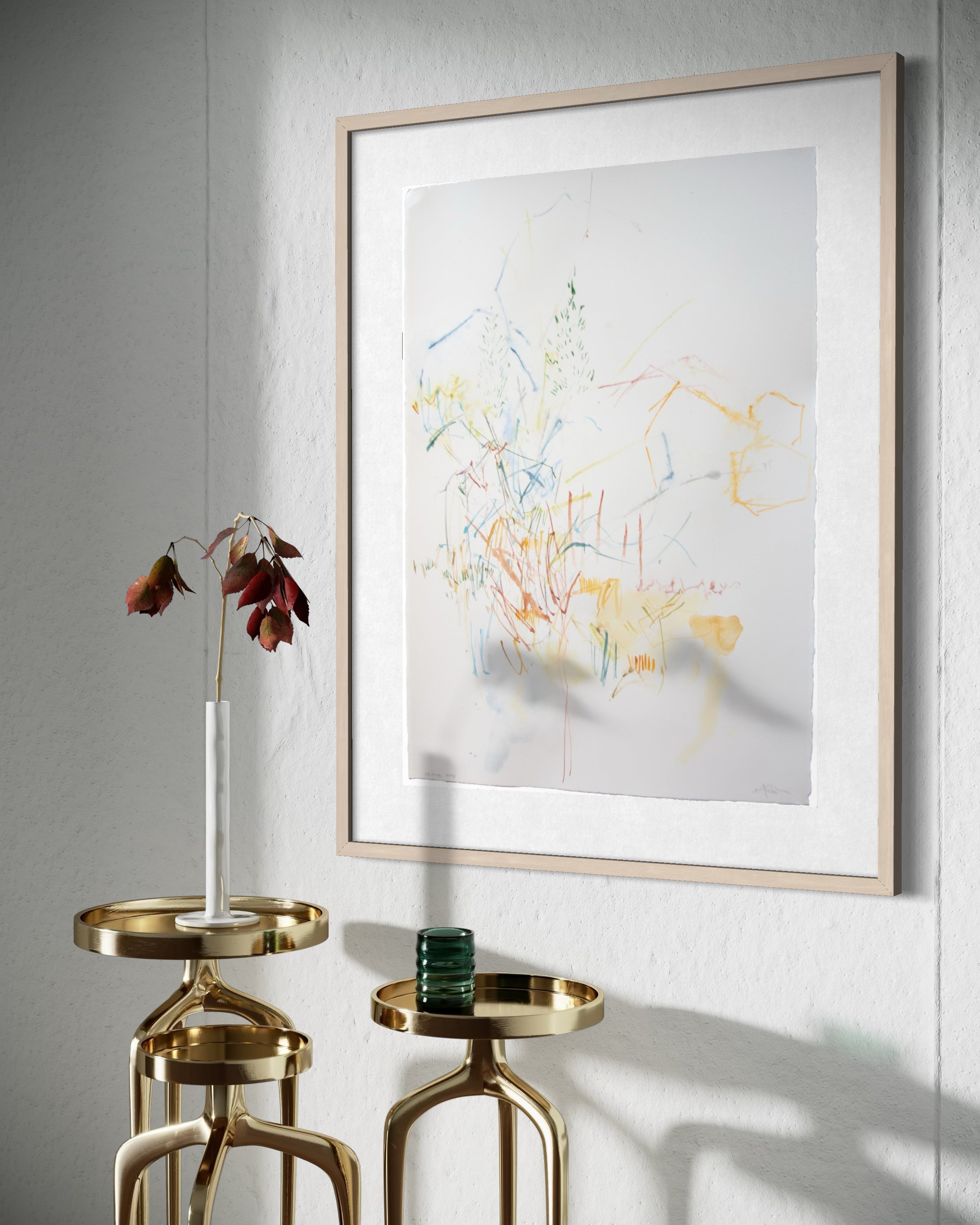 A framed abstract art print with delicate multicolored brushstrokes on a white background, styled in a minimalist interior setting with gold side tables and a dried flower vase.