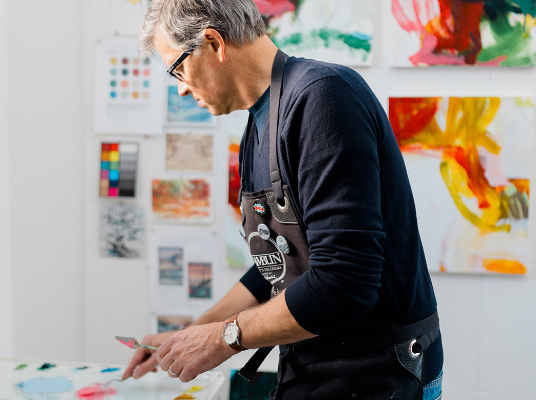 Artist Michael Rich wearing a dark navy long-sleeve shirt and a brown artist's apron, painting at a table in an art studio with colorful canvases on the wall.
