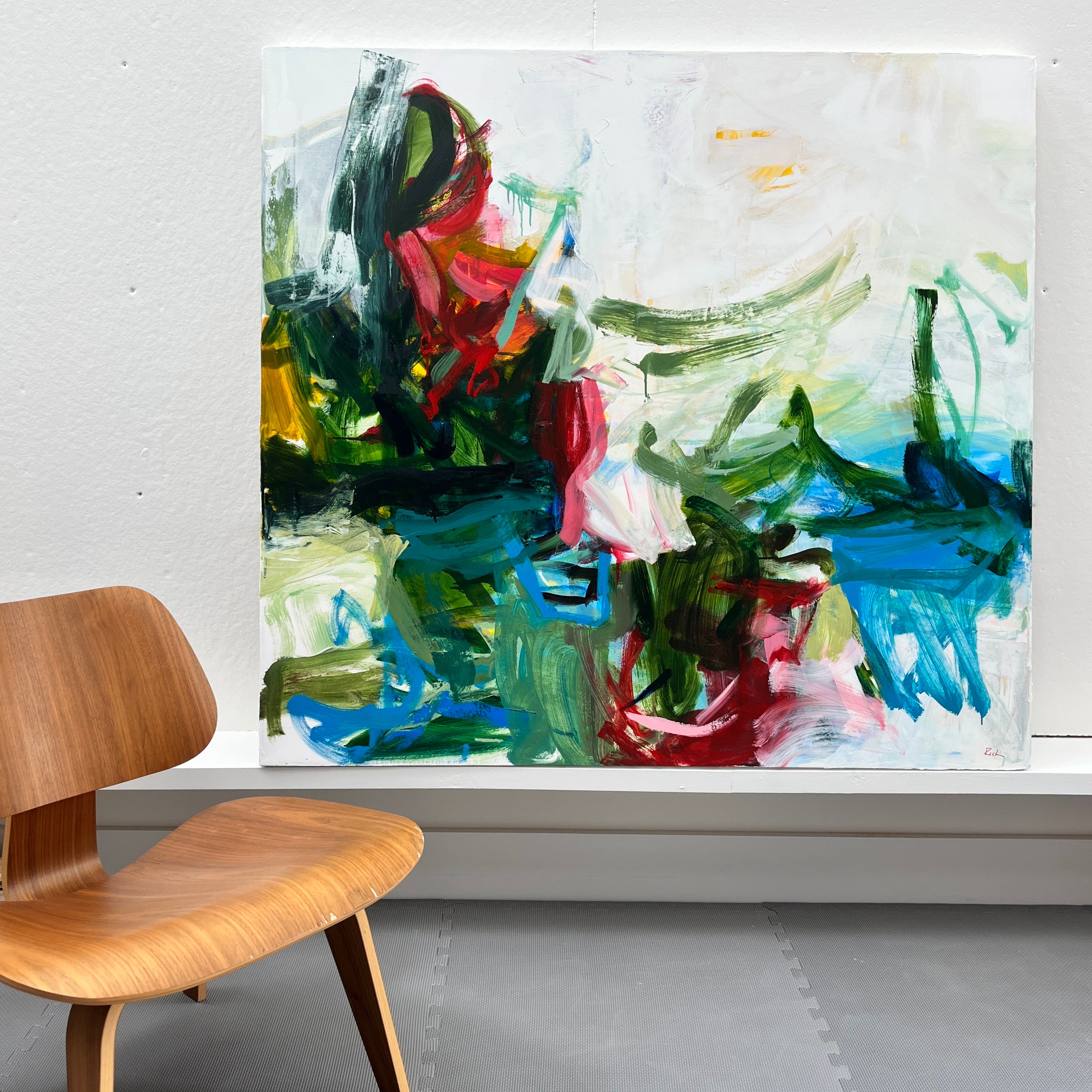 An abstract canvas painting with expressive brushstrokes in green, blue, red, white, and black, displayed on a white wall above a white ledge, with a wooden chair partially visible in the foreground.