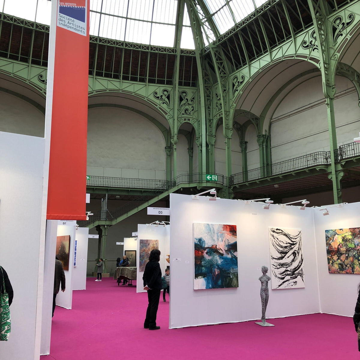 An indoor art fair or exhibition space with white display walls showcasing colorful abstract paintings, a metallic sculpture, and a person standing nearby. The venue has a bright pink carpet and a large industrial hall with green metal arches and a glass roof.
