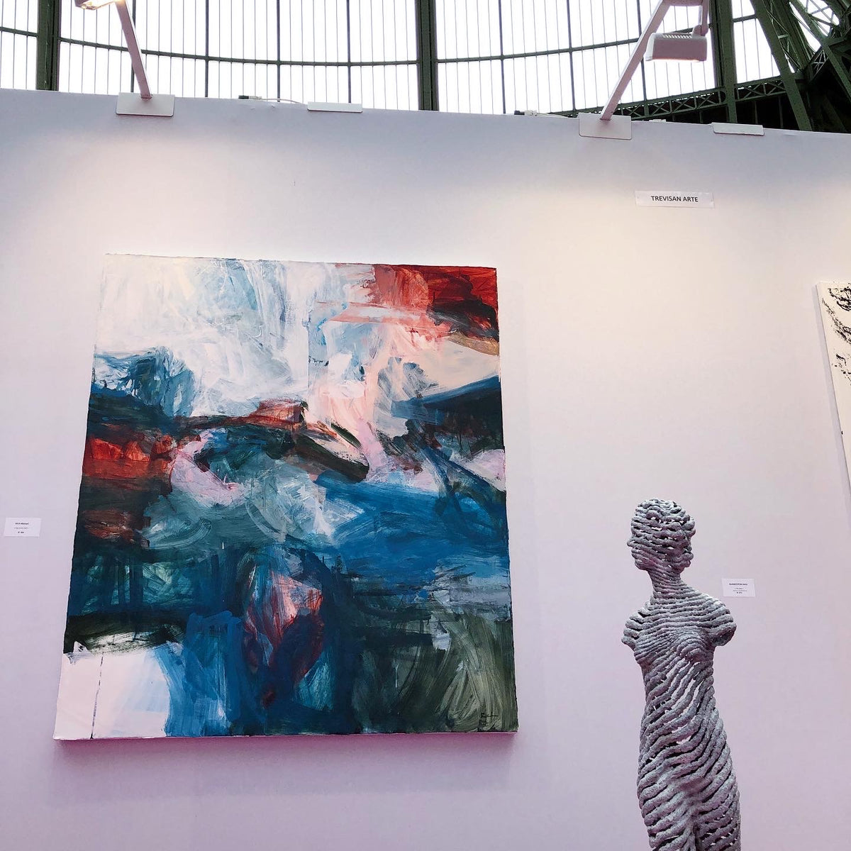An abstract painting by Michael Rich with blue, white, red, and black brushstrokes displayed on a white gallery wall next to a gray textured female-form sculpture.