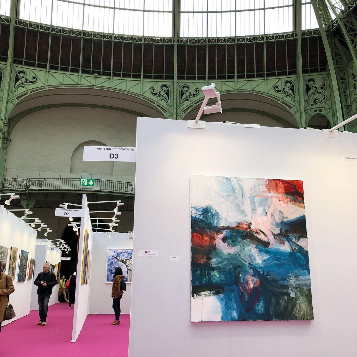 An abstract painting by Michael Rich with blue, red, white, and black brushstrokes displayed on a white wall at an art fair inside a large industrial hall with green metal architecture.