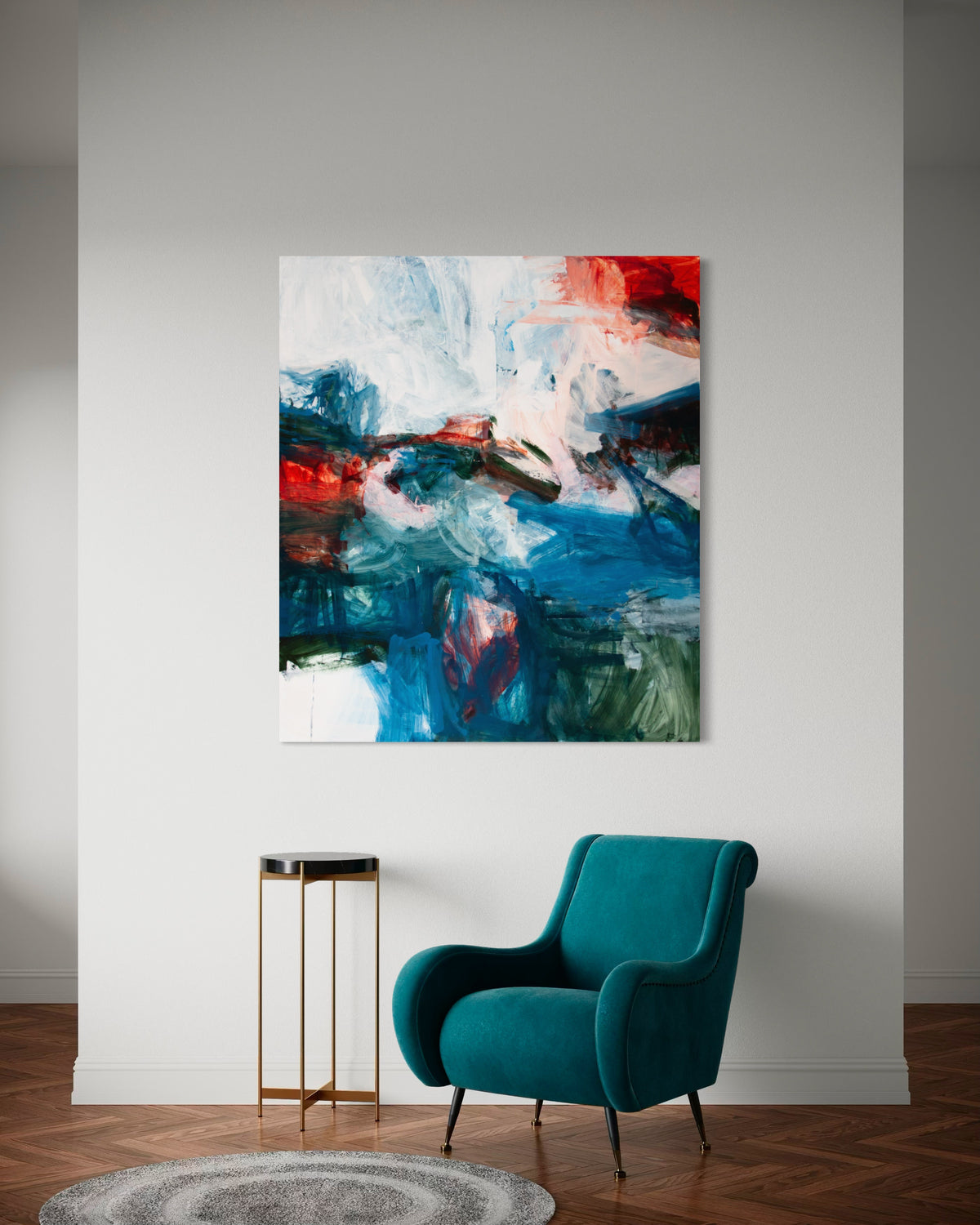 An abstract canvas painting by Michael Rich with blue, red, white, and black brushstrokes displayed on a light gray wall above a teal armchair and a small gold side table.