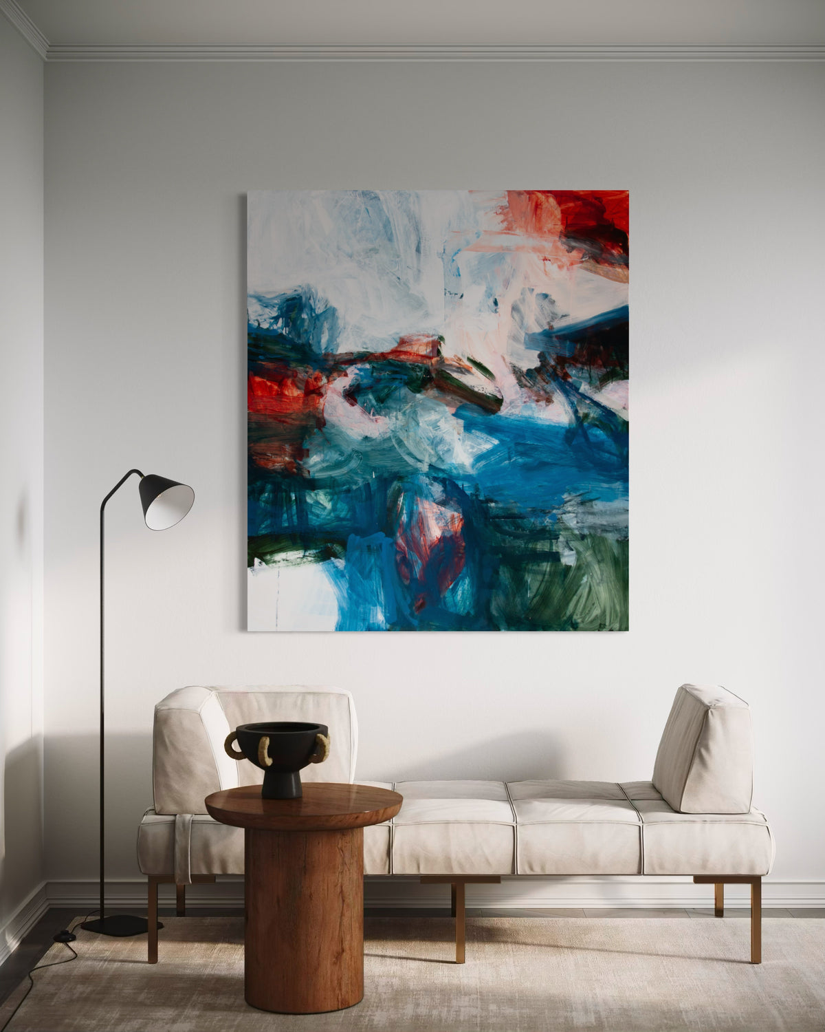 An abstract canvas painting by Michael Rich with blue, white, red, and green brushstrokes, displayed on a light gray wall above a beige sofa in a modern living room setting.