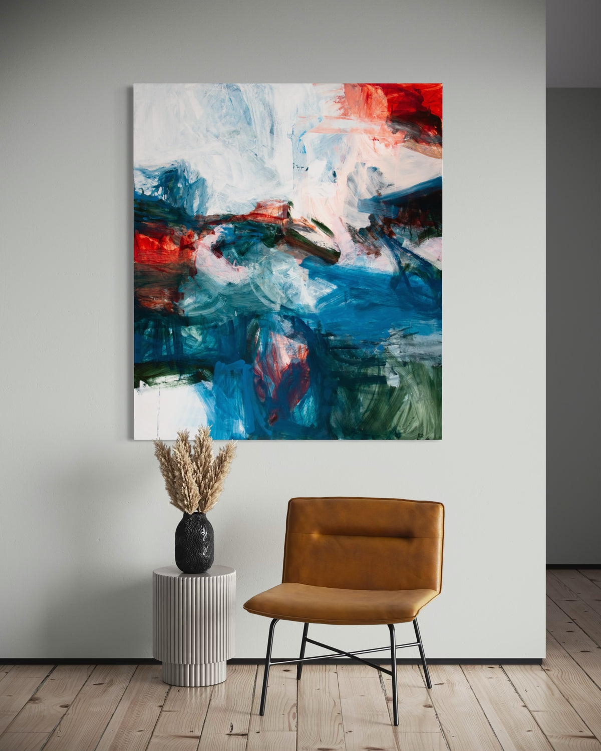 An abstract canvas painting by Michael Rich with vibrant blue, red, white, and green brushstrokes, displayed on a light gray wall in a modern room setting above a brown leather chair and a small side table.