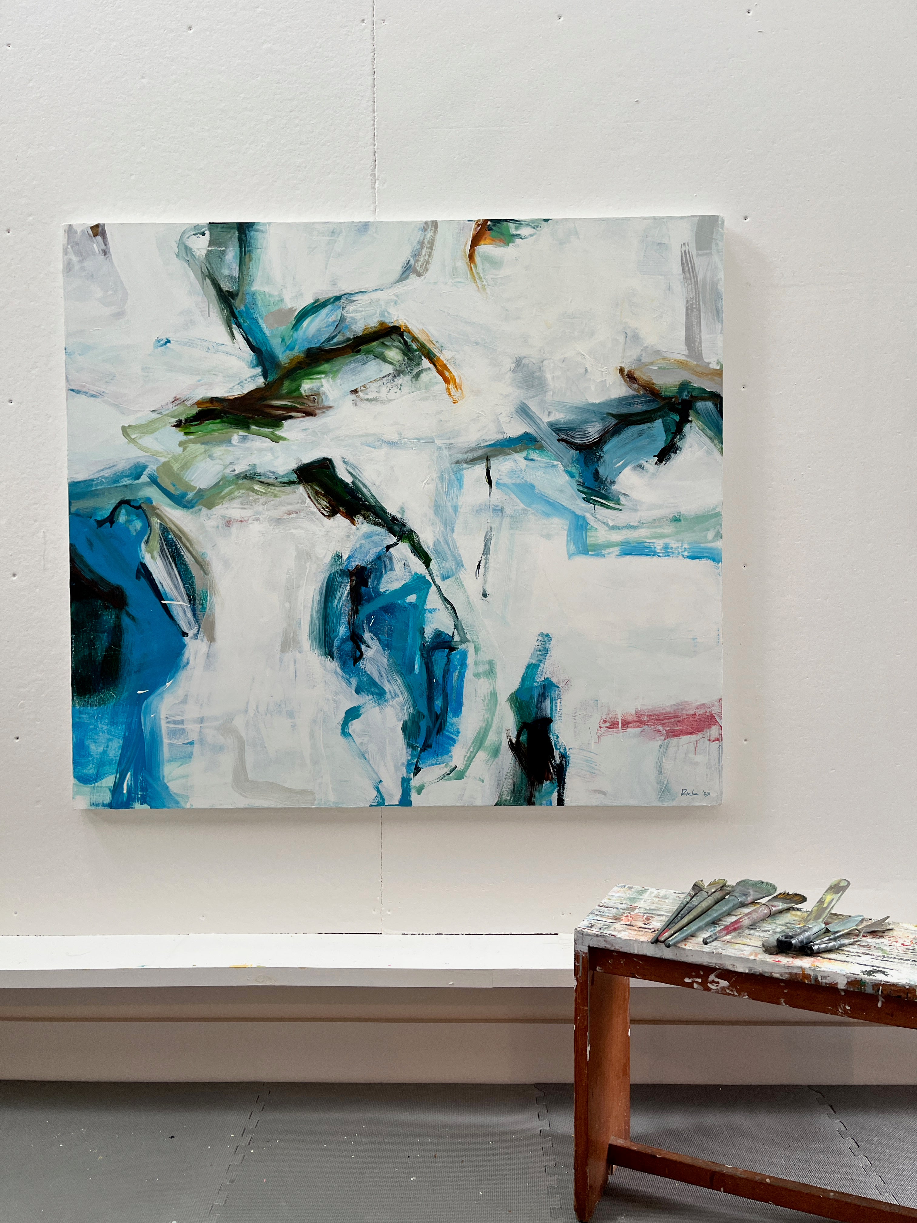 An abstract canvas painting with blue, green, black, and white brushstrokes hanging on a white wall, with a wooden stool holding paintbrushes in the foreground.