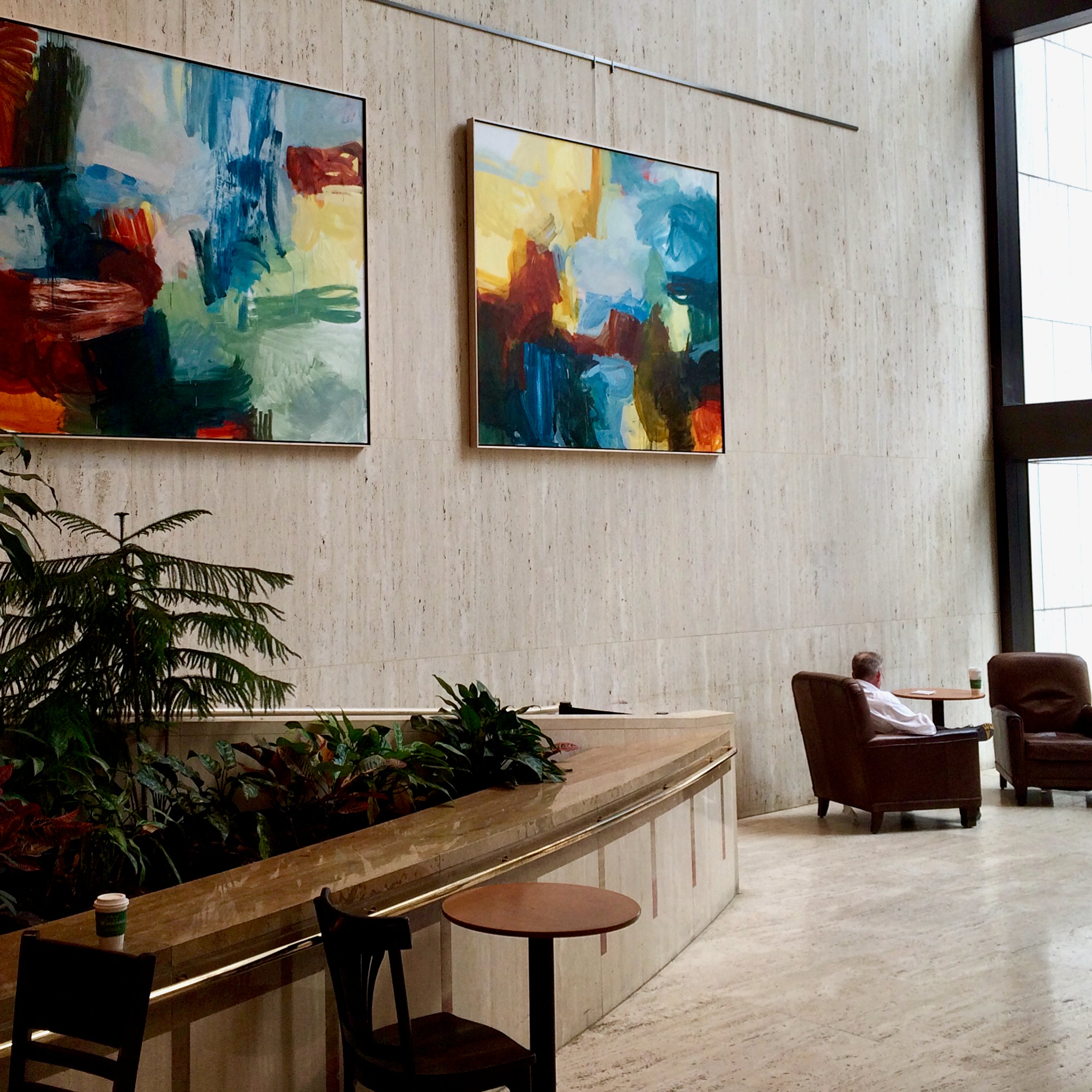 Two large abstract canvas paintings with vibrant blue, green, yellow, red, and orange brushstrokes hang on a textured beige wall in a modern, sunlit lobby. Below the paintings is a long wooden planter bed filled with green plants.