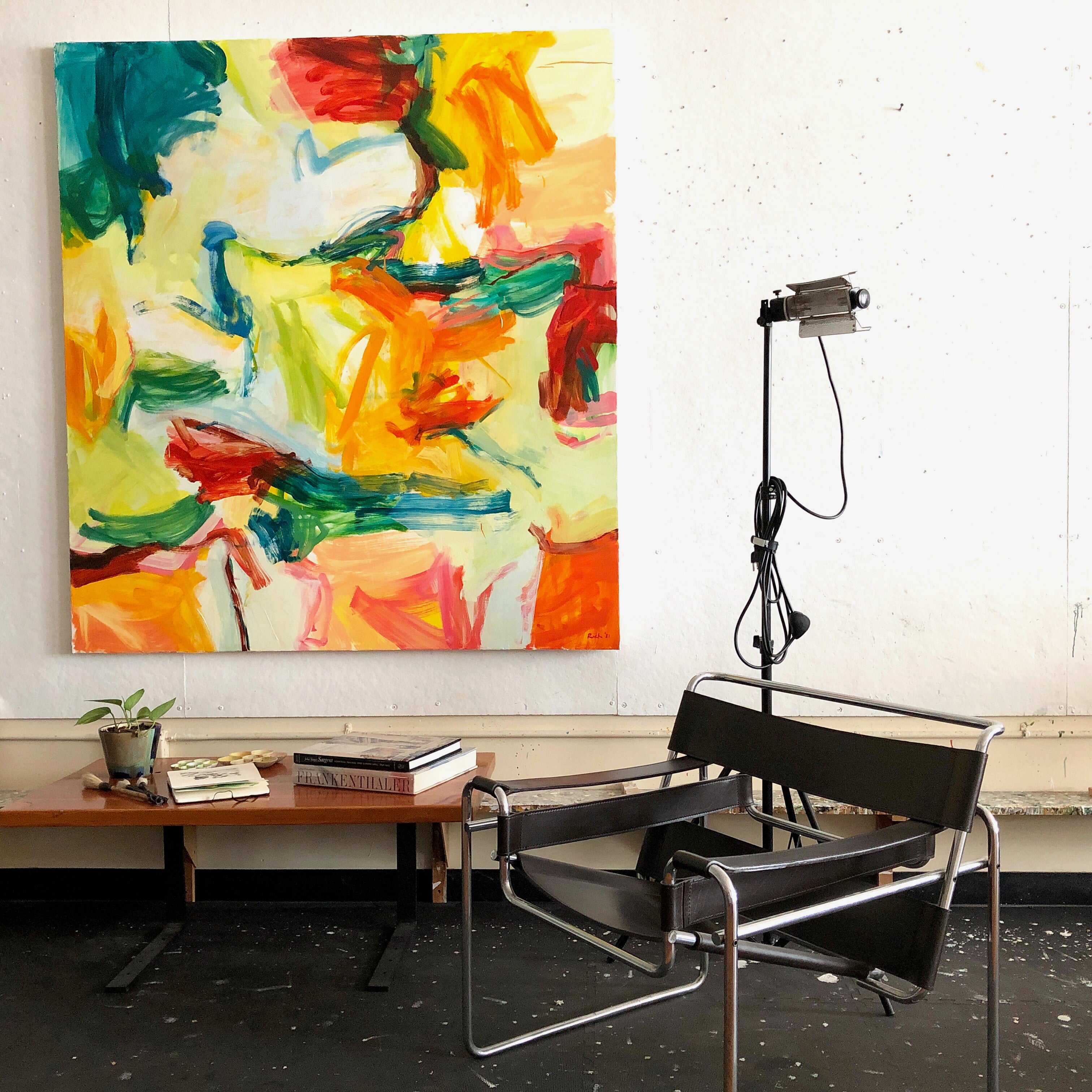 A large abstract canvas painting with bold strokes of yellow, orange, red, green, and blue hangs on a white wall in a studio-like room. In front of the painting is a black leather and chrome chair, with a small table, books, and a plant below.