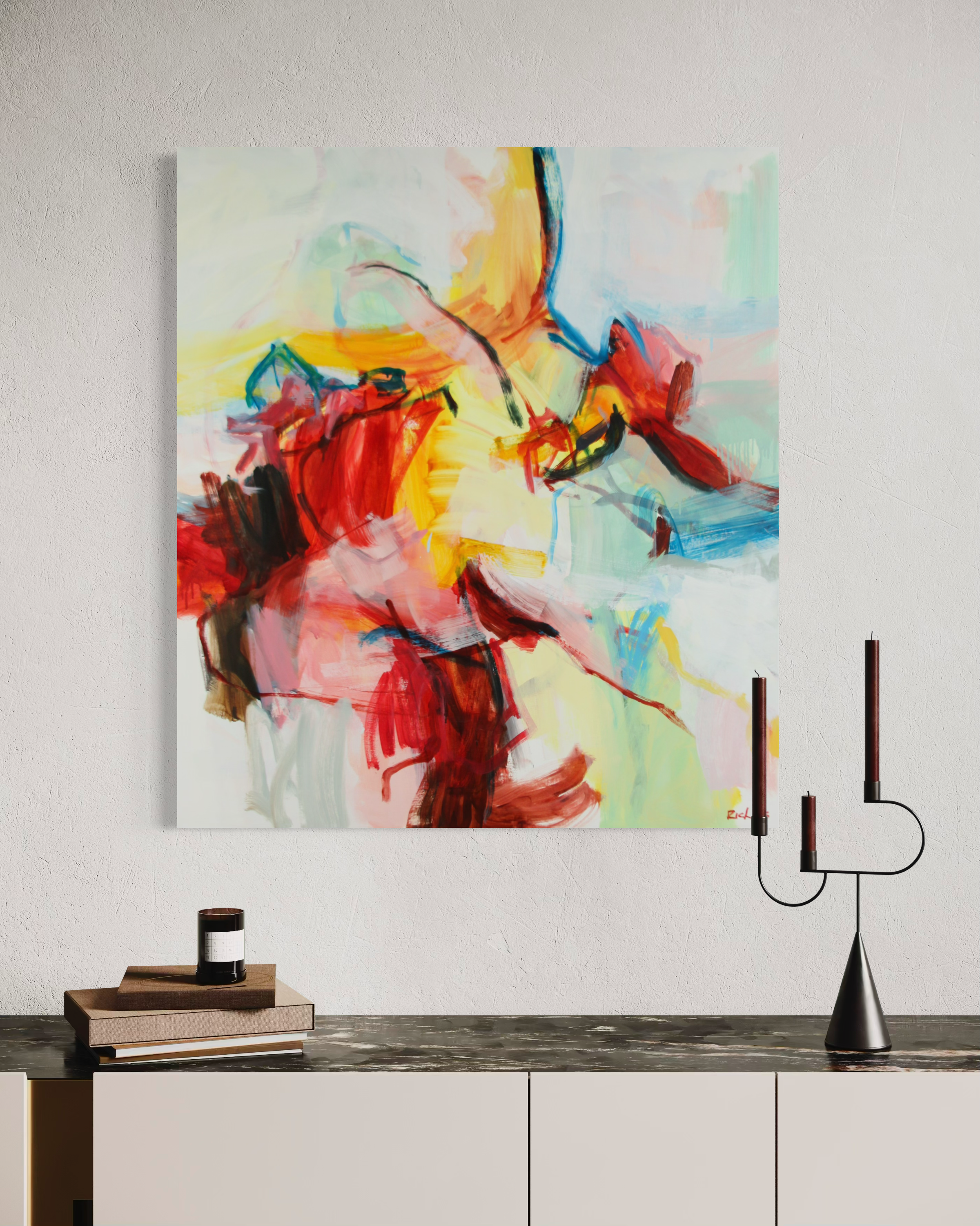 Abstract canvas wall art featuring expressive brushstrokes in red, yellow, blue, green, and black on a white background, displayed in a styled interior above a console table with books and a candle holder.