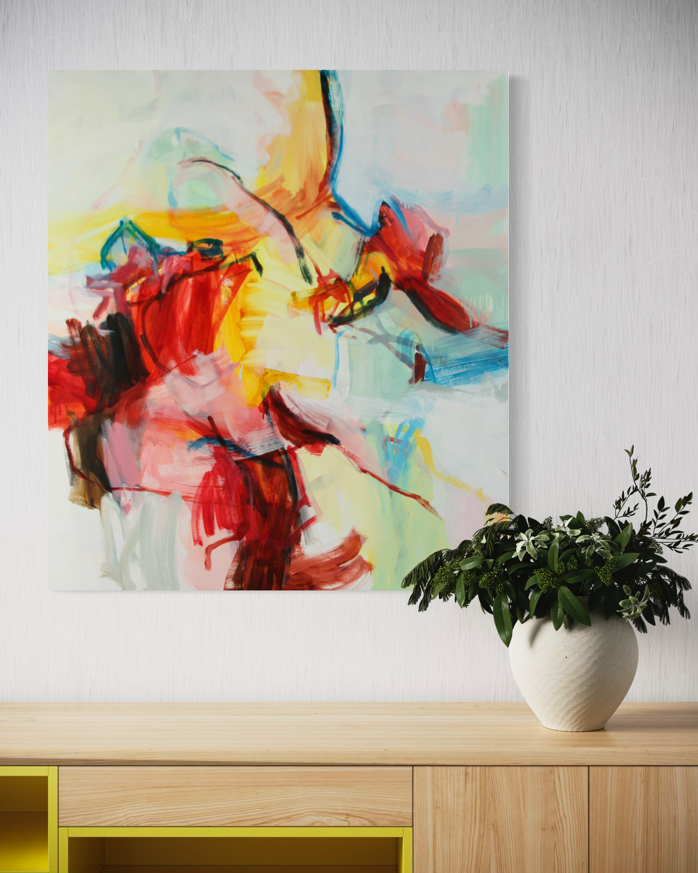 An abstract canvas painting with bold brushstrokes of red, yellow, white, blue, and black, displayed on a white wall above a light wood console table with a potted green plant in a white vase.