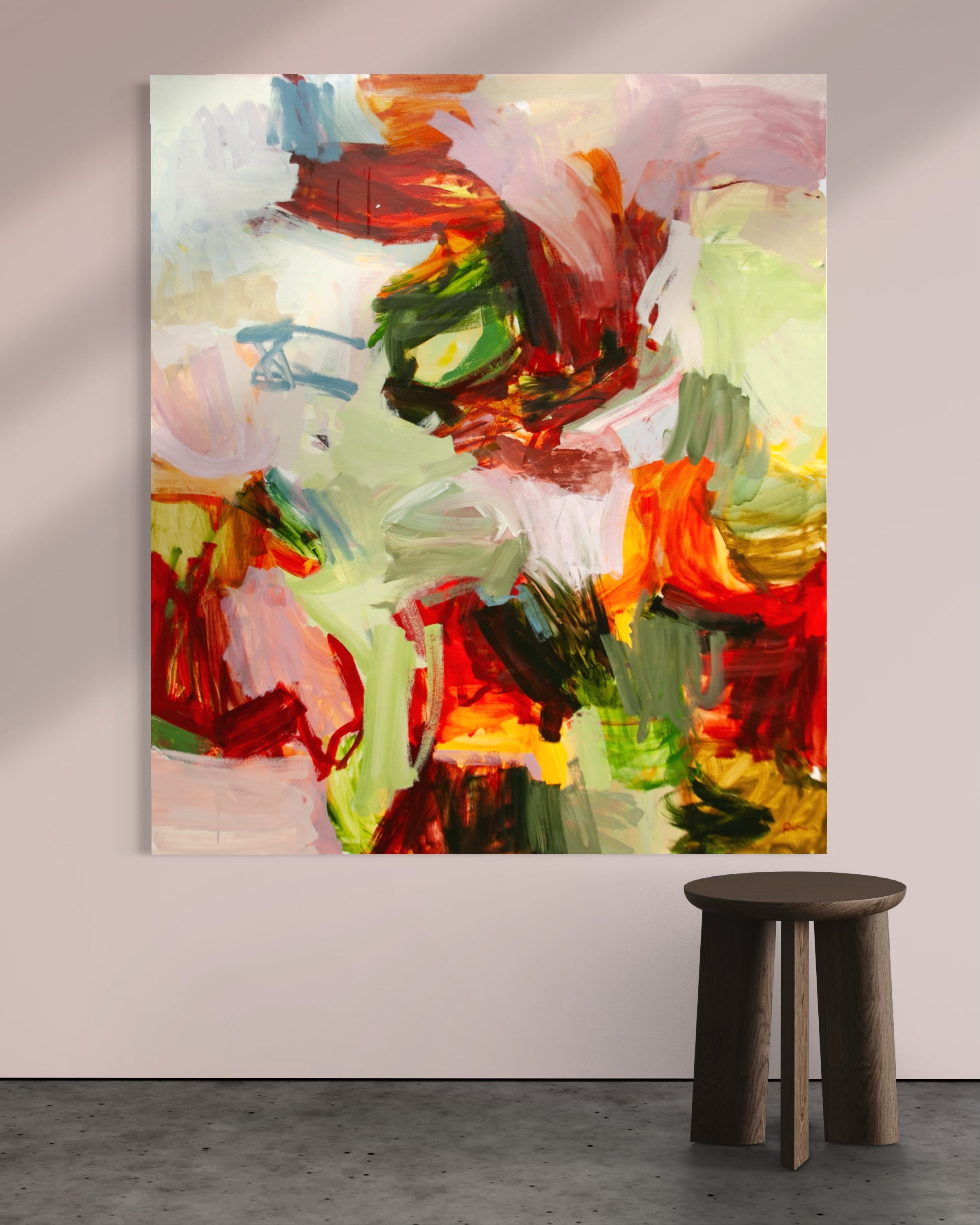 An abstract canvas painting with expressive brushstrokes in red, green, yellow, pink, and white, displayed on a light beige wall above a dark brown wooden stool.