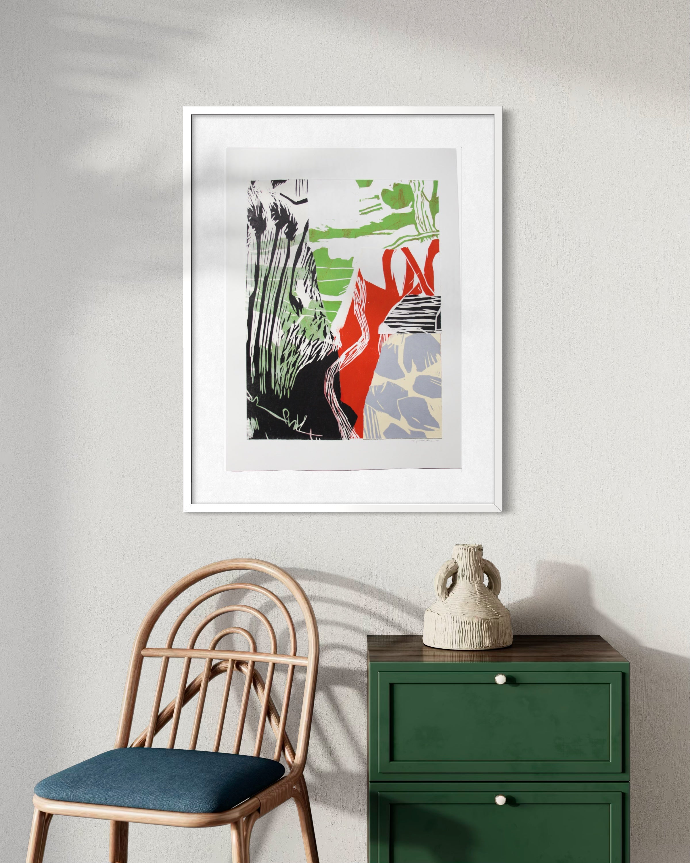 A framed abstract art print with green, red, black, and gray brushstrokes on a white mat, hanging on a light gray wall above a green cabinet and a wooden chair.