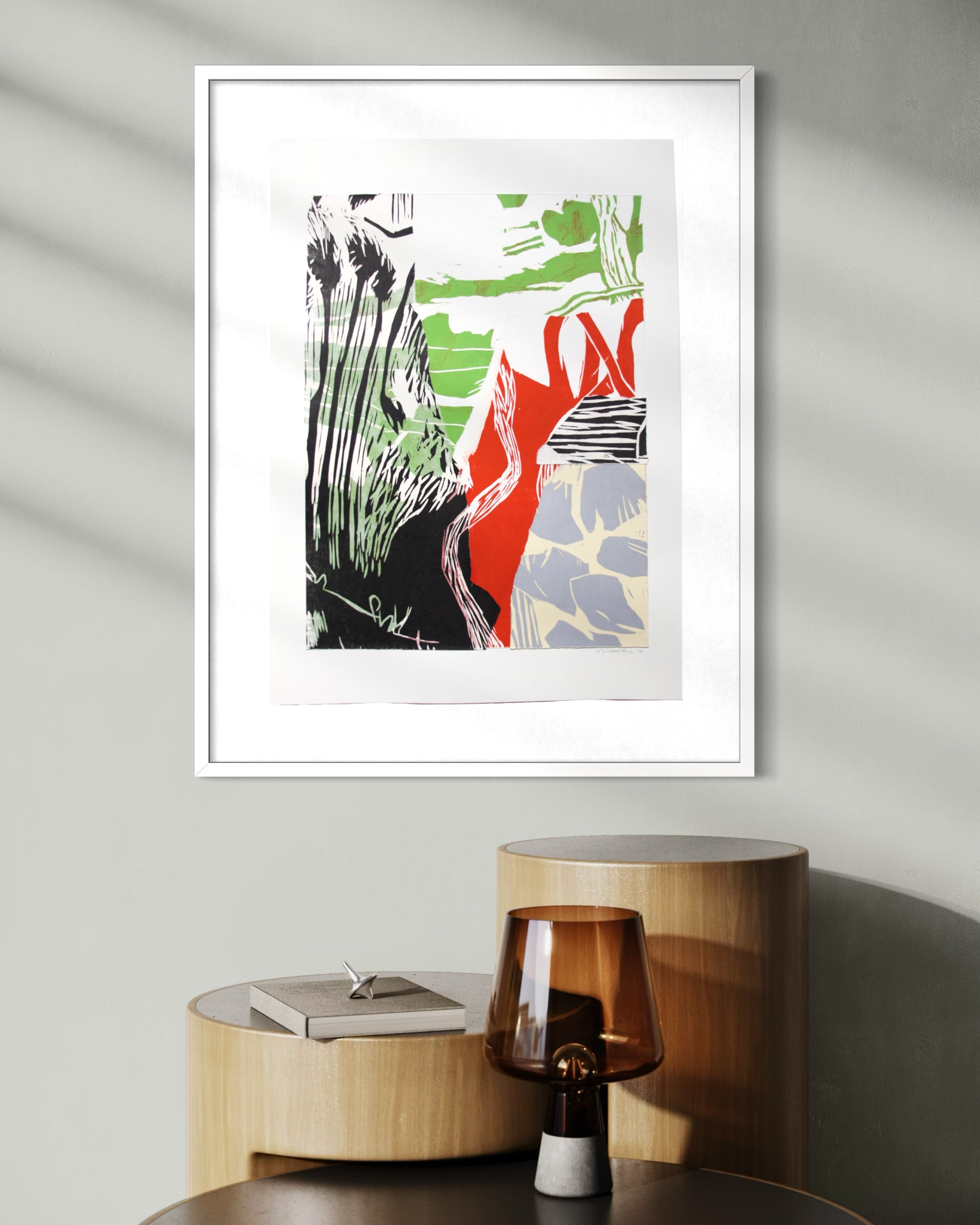 A framed abstract art print with bold black, green, red, and gray brushstrokes on a white background, displayed on a wall above a wooden side table with a glass lamp.