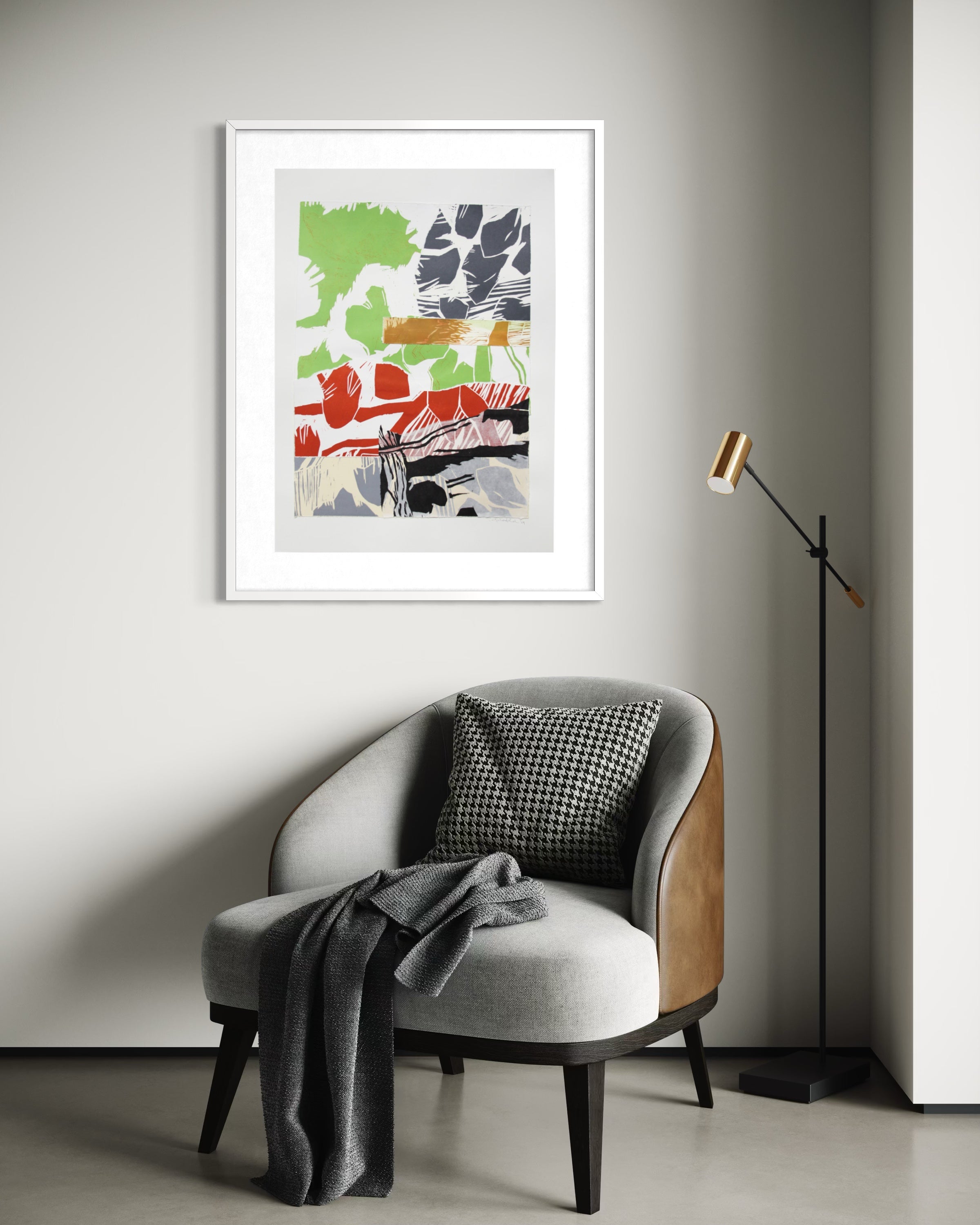A framed abstract art print with green, red, gray, and orange brushstrokes on a light gray background, displayed on a gray wall above a modern gray armchair with a black-and-white checkered pillow and a draped gray throw blanket. A black floor lamp stands to the right.