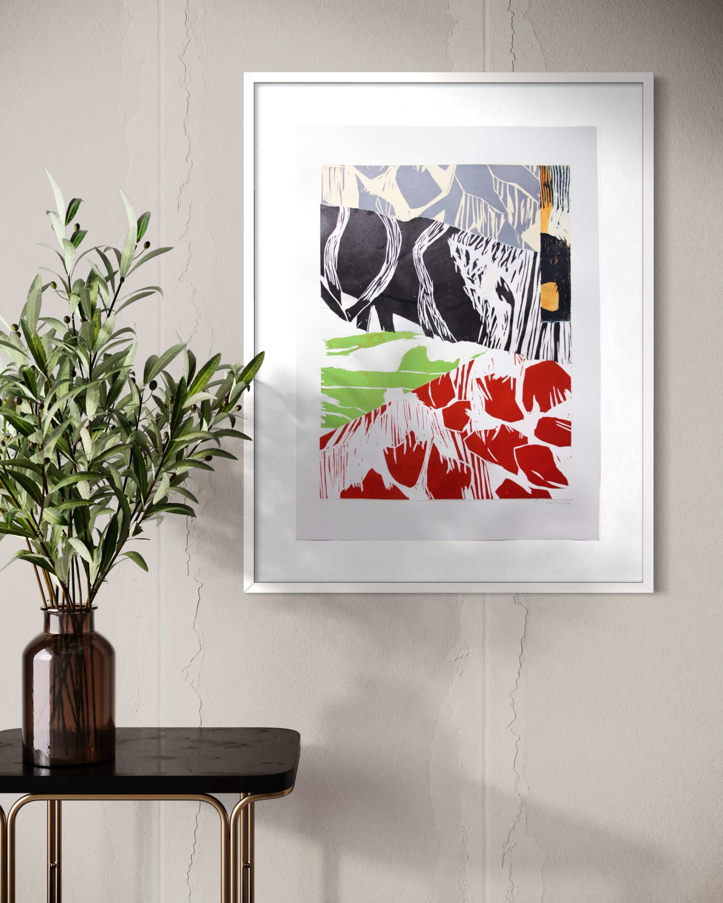 A framed abstract art print with black, white, red, and green brushstrokes, displayed on a light gray wall above a side table with a green plant in a dark glass vase.