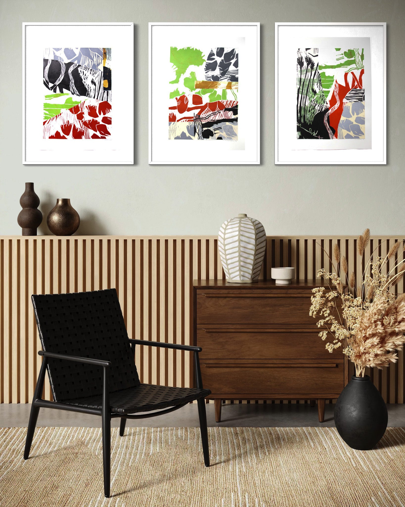Three framed abstract art prints hang on a light gray wall above a slatted wood panel, with a black armchair, wooden cabinet, ceramic vase, and dried floral arrangement in a styled interior setting.