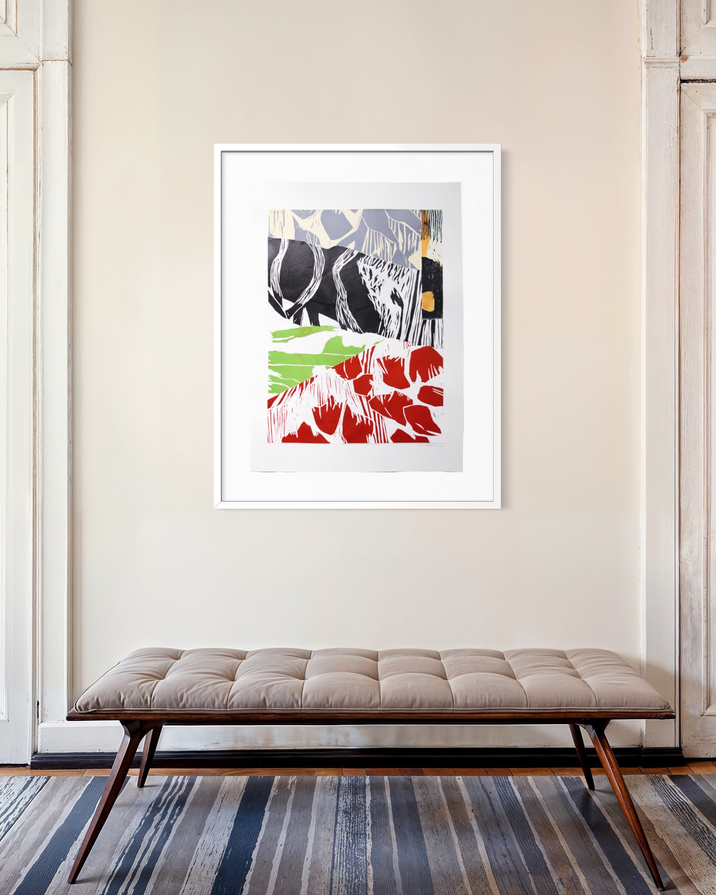 A framed abstract art print with red, green, black, and gray brushstrokes hangs on a beige wall above a tufted bench in a styled interior setting.
