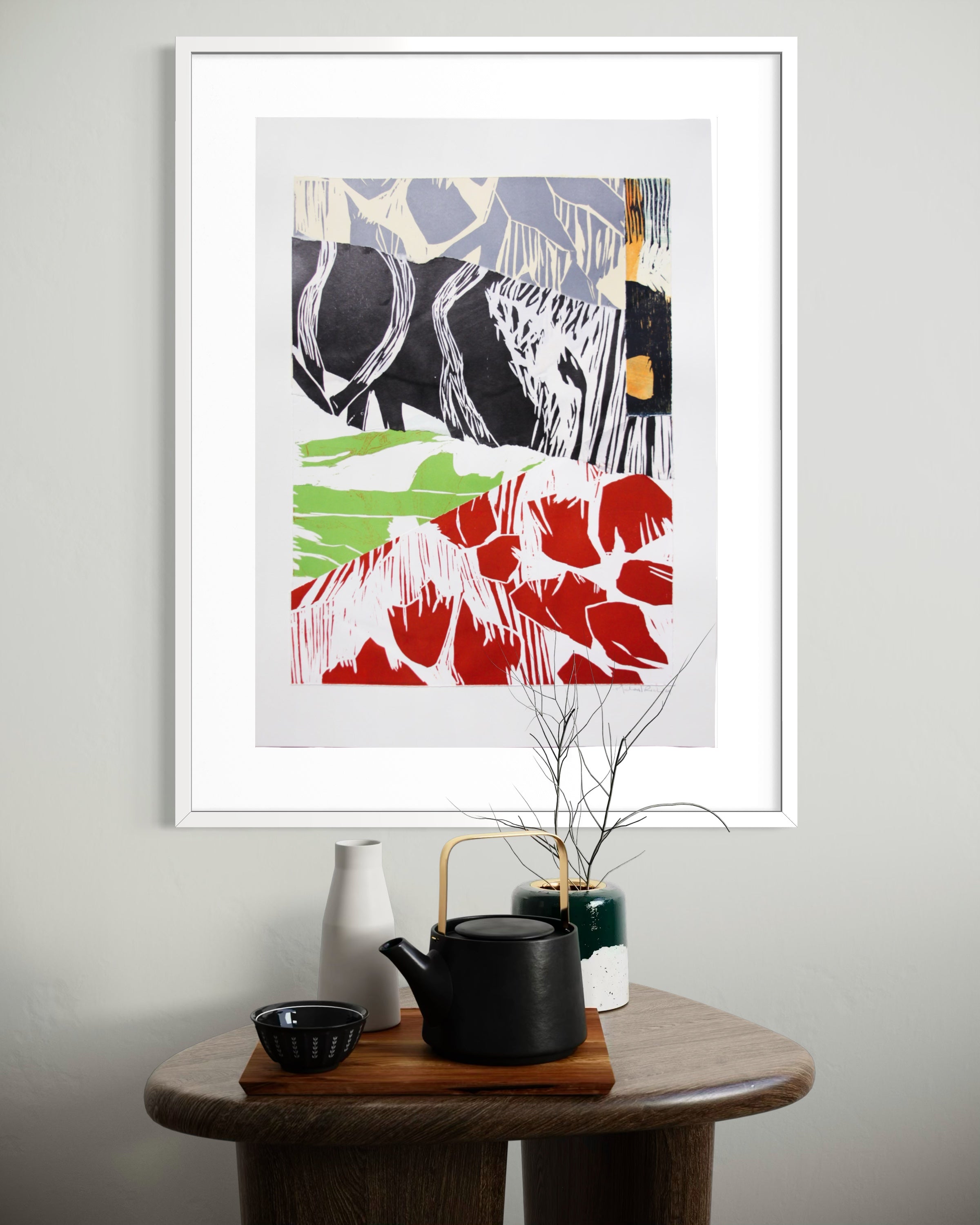 A framed abstract art print with bold black, red, green, and gray brushstrokes, displayed on a light gray wall above a wooden side table with a black teapot, white vase, and small green pot.