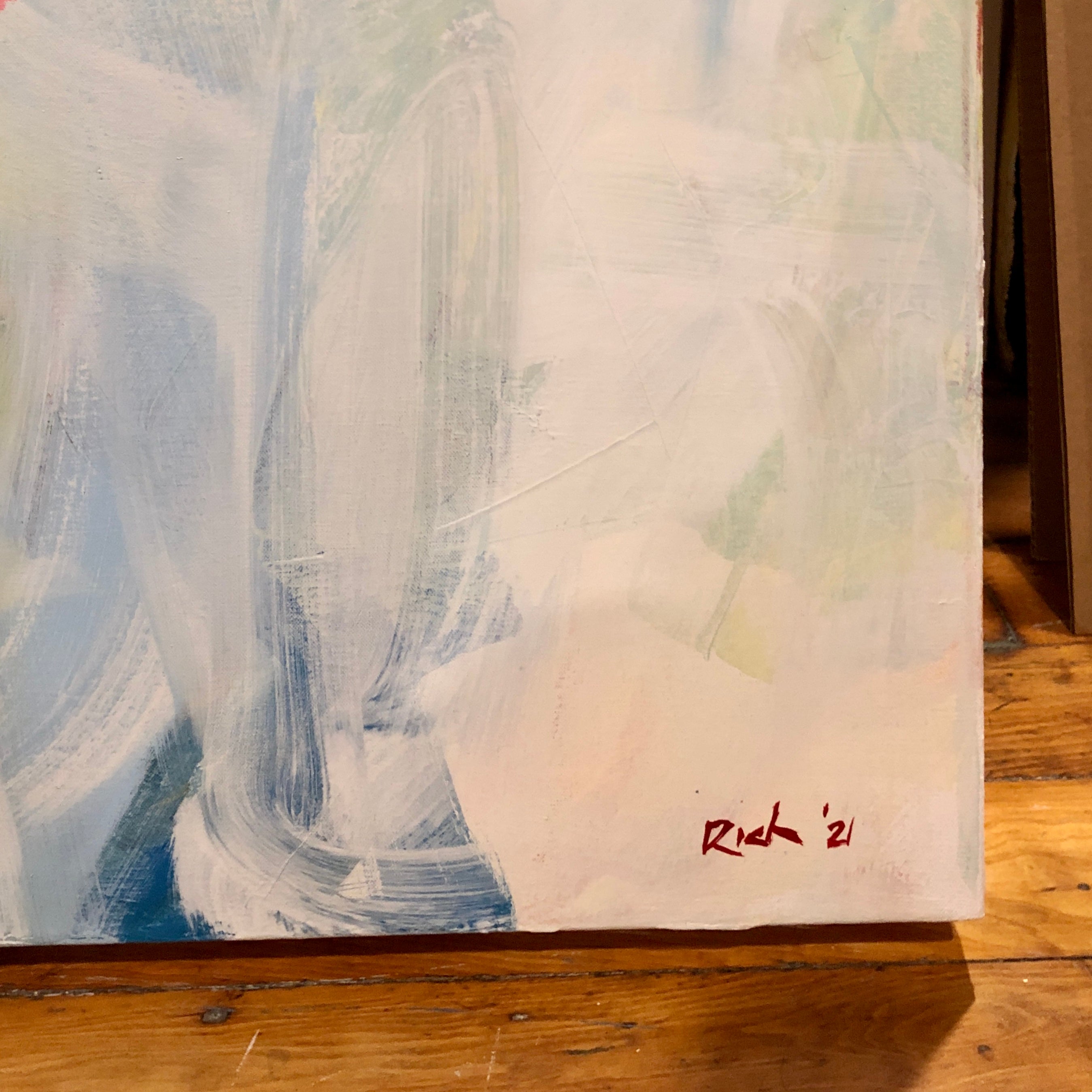 An abstract canvas painting with textured brushstrokes in white, light blue, and gray, featuring a small red signature reading 'Rich '21' on the bottom right, displayed on a wooden floor.