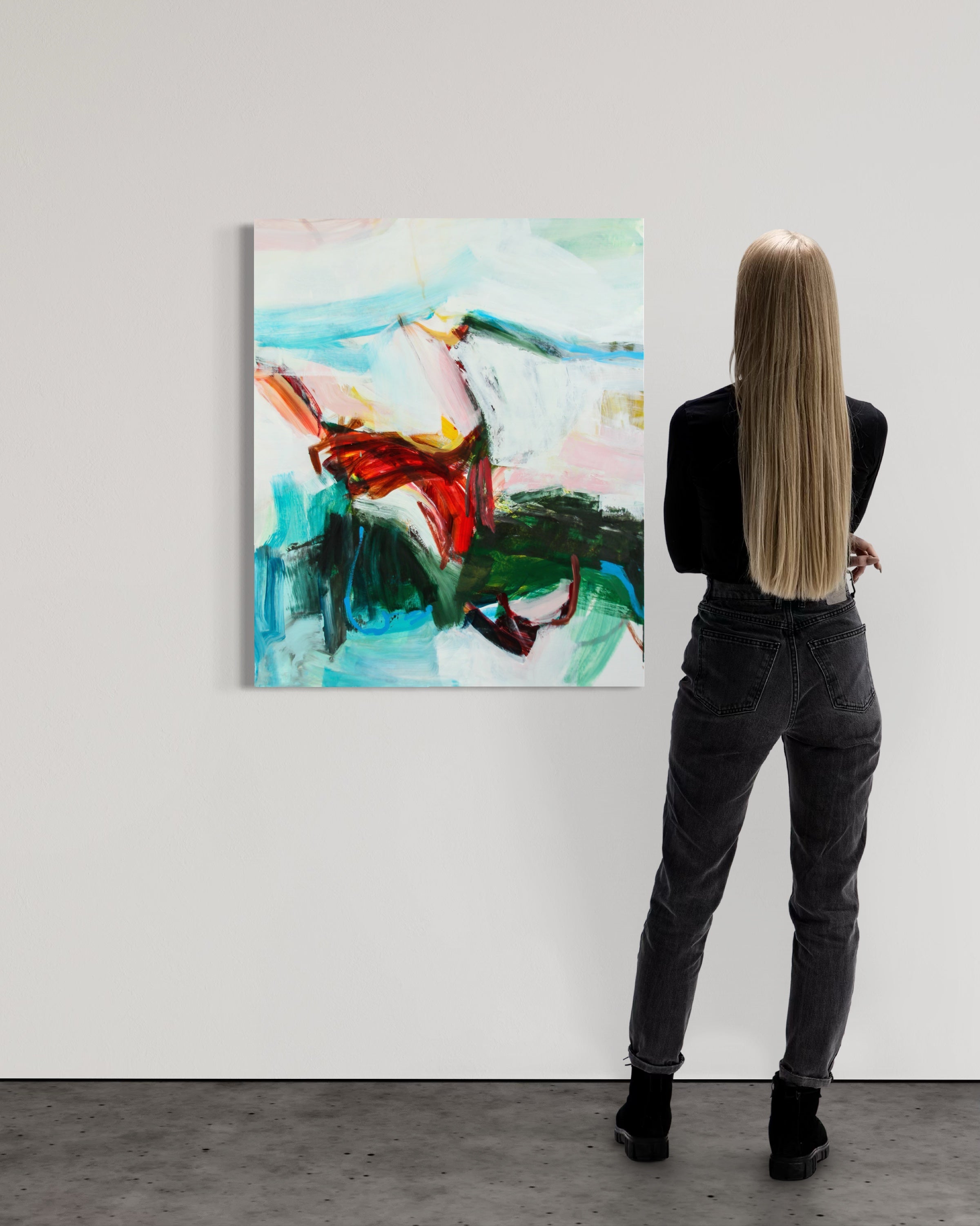 A woman with long blonde hair stands with her back to the camera, looking at an abstract painting hanging on a white gallery wall. The painting features expressive brushstrokes in light blue, white, green, and red.