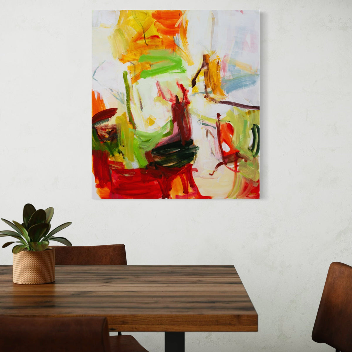 An abstract canvas painting by Michael Rich with vibrant brushstrokes of red, orange, yellow, green, and white, displayed on a textured white wall above a wooden dining table with chairs and a small potted plant.