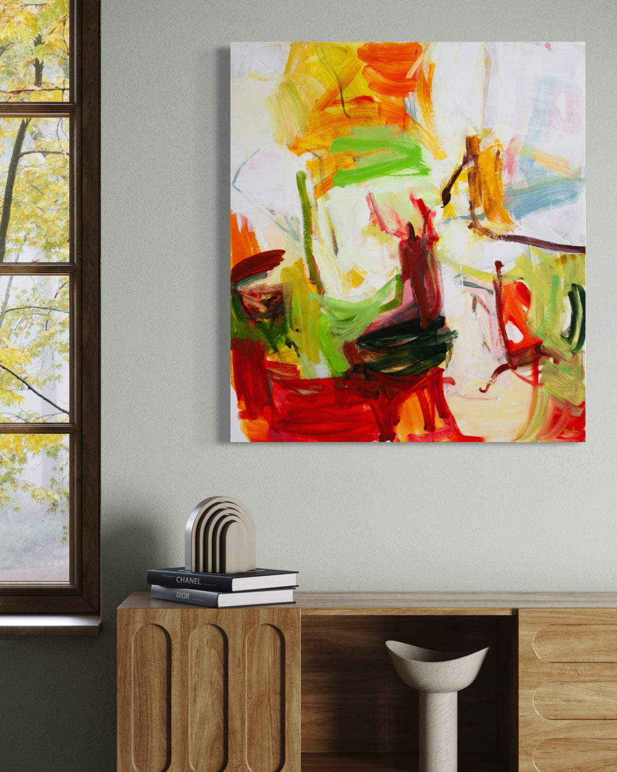 An abstract canvas painting by Michael Rich with vibrant red, orange, green, yellow, and white brushstrokes, displayed on a light gray wall above a wooden console table in a modern room setting.