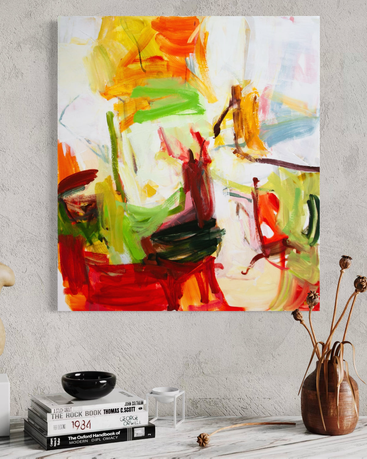 An abstract canvas painting by Michael Rich with expressive brushstrokes in red, orange, green, yellow, and white, displayed on a light gray textured wall above a styled tabletop with books and dried flowers.
