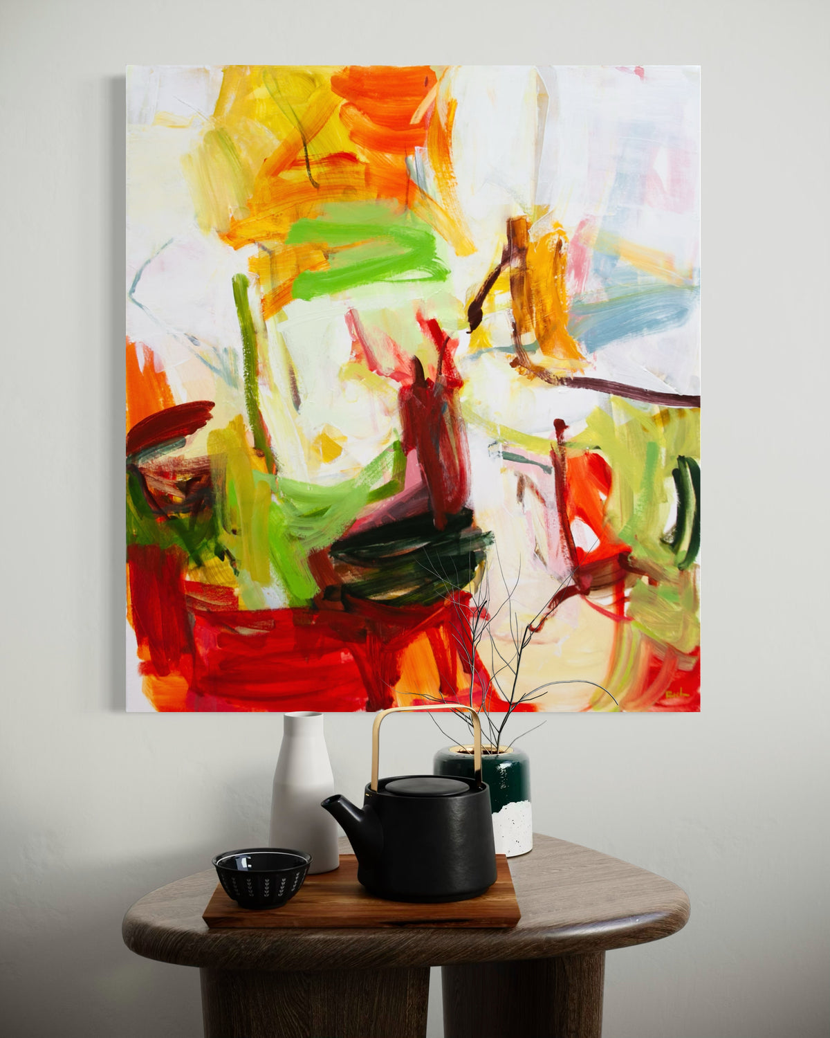 An abstract canvas painting by Michael Rich with expressive brushstrokes in red, orange, yellow, green, and white, displayed on a light gray wall above a wooden side table with a black teapot and small vases.