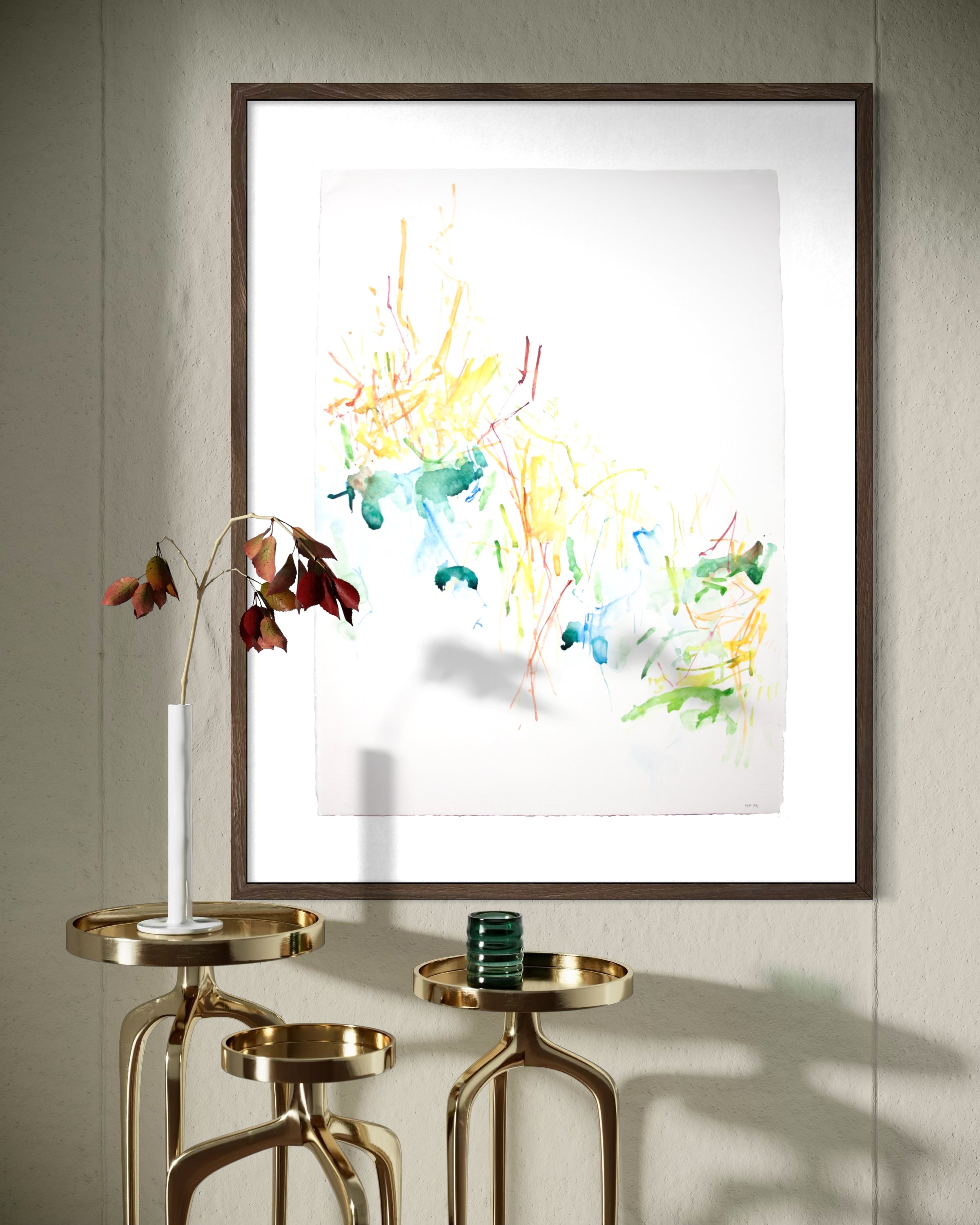 A framed abstract watercolor art print with colorful brushstrokes on a white background, hanging on a light gray paneled wall above two gold side tables with candles.