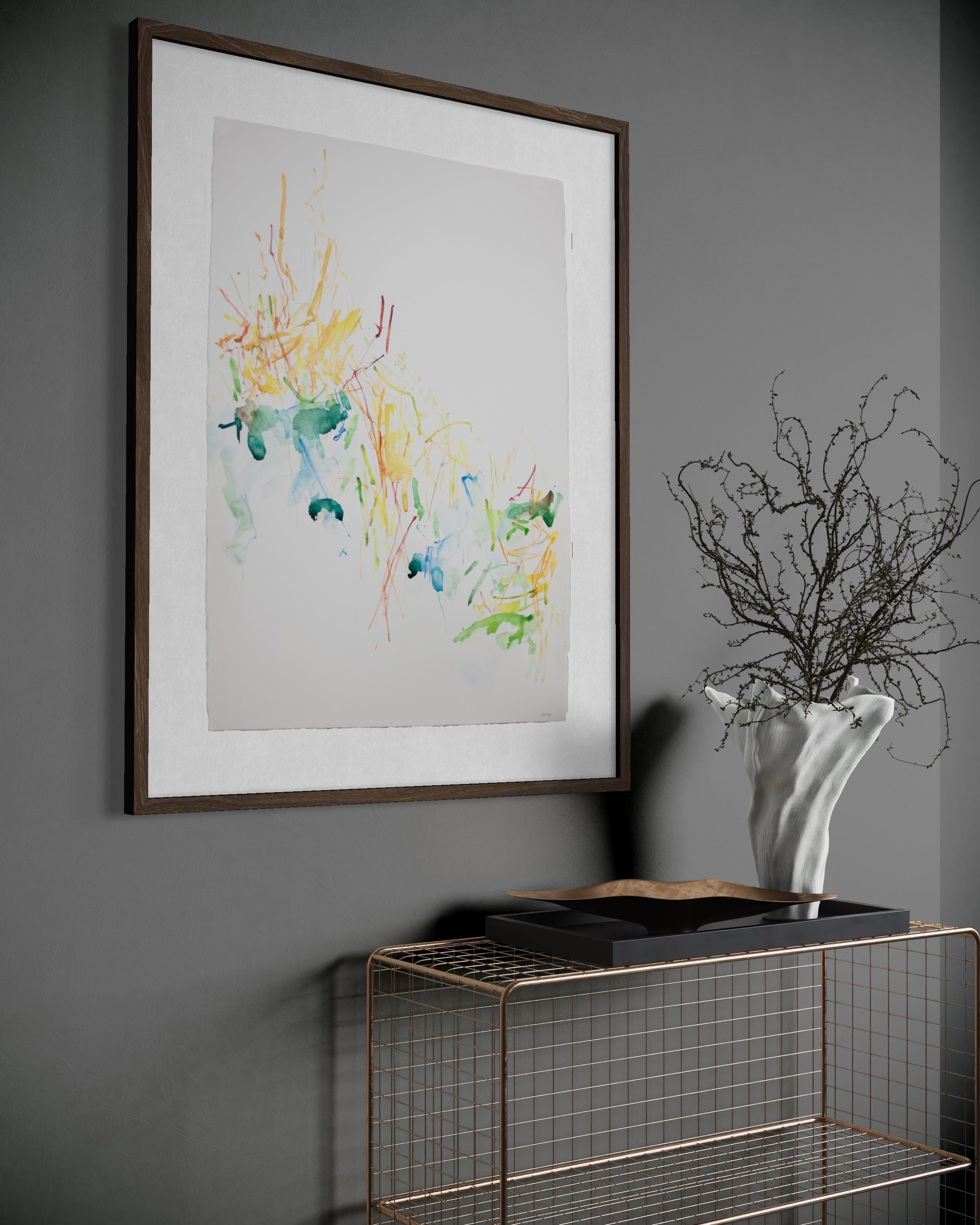 A framed abstract art print with colorful watercolor-like splashes on a white background, hung on a dark gray wall above a wire console table with decorative branches in a white vase.