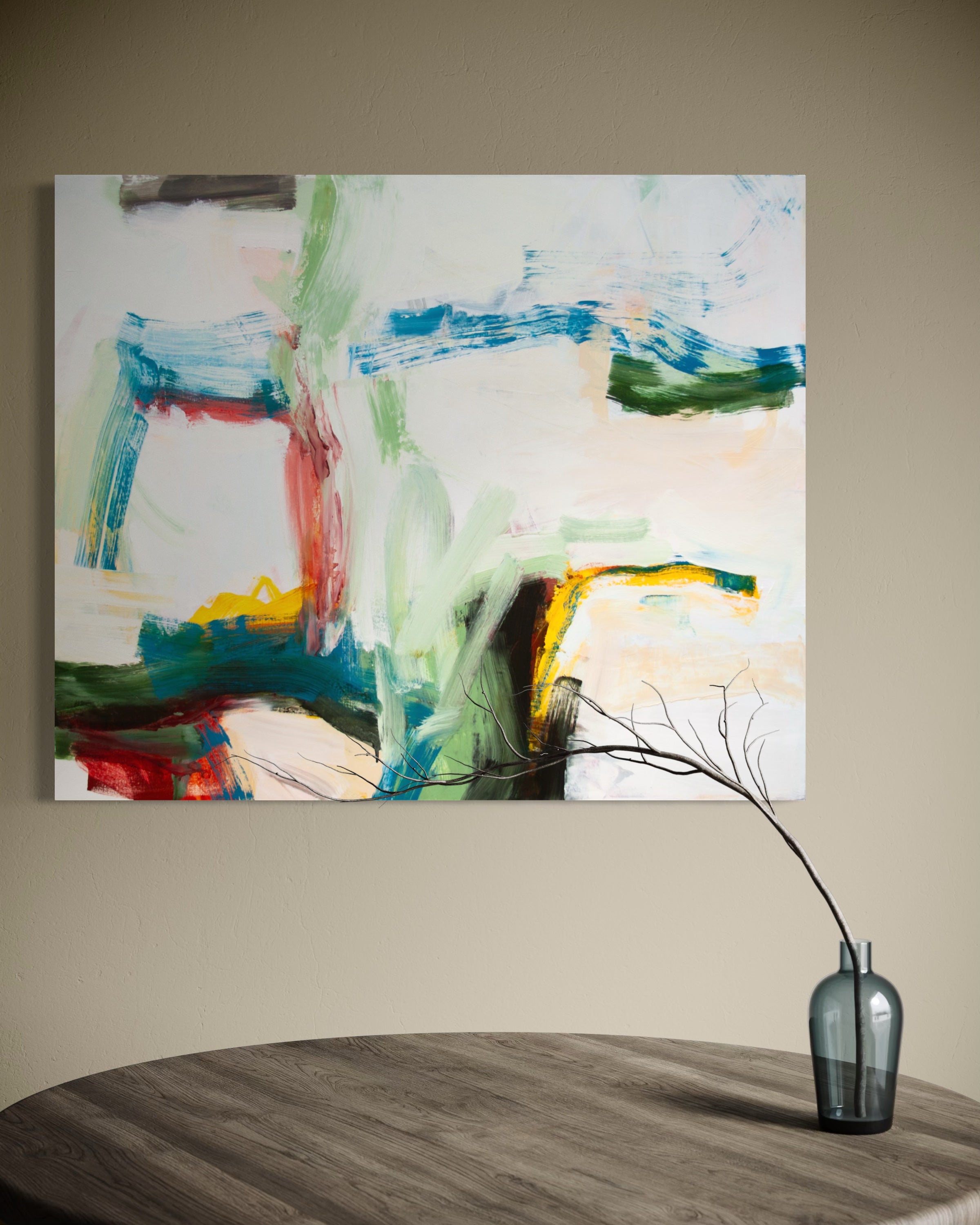 An abstract canvas painting with blue, green, red, yellow, and black brushstrokes on a white background, displayed on a beige wall above a dark wood table with a glass vase.