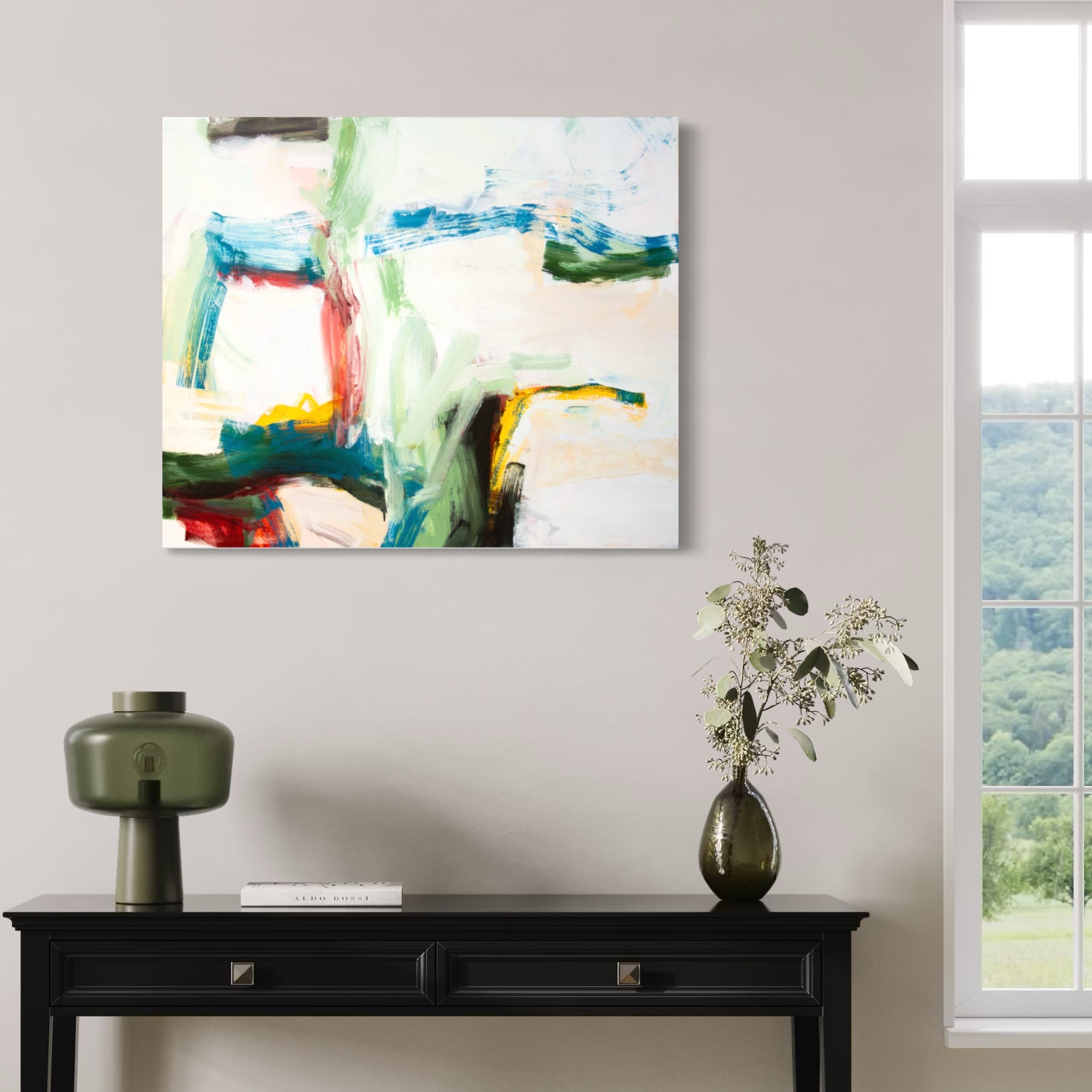 An abstract canvas wall art print with green, blue, red, and white brushstrokes, displayed in a styled room above a black console table with a lamp and a vase of flowers.