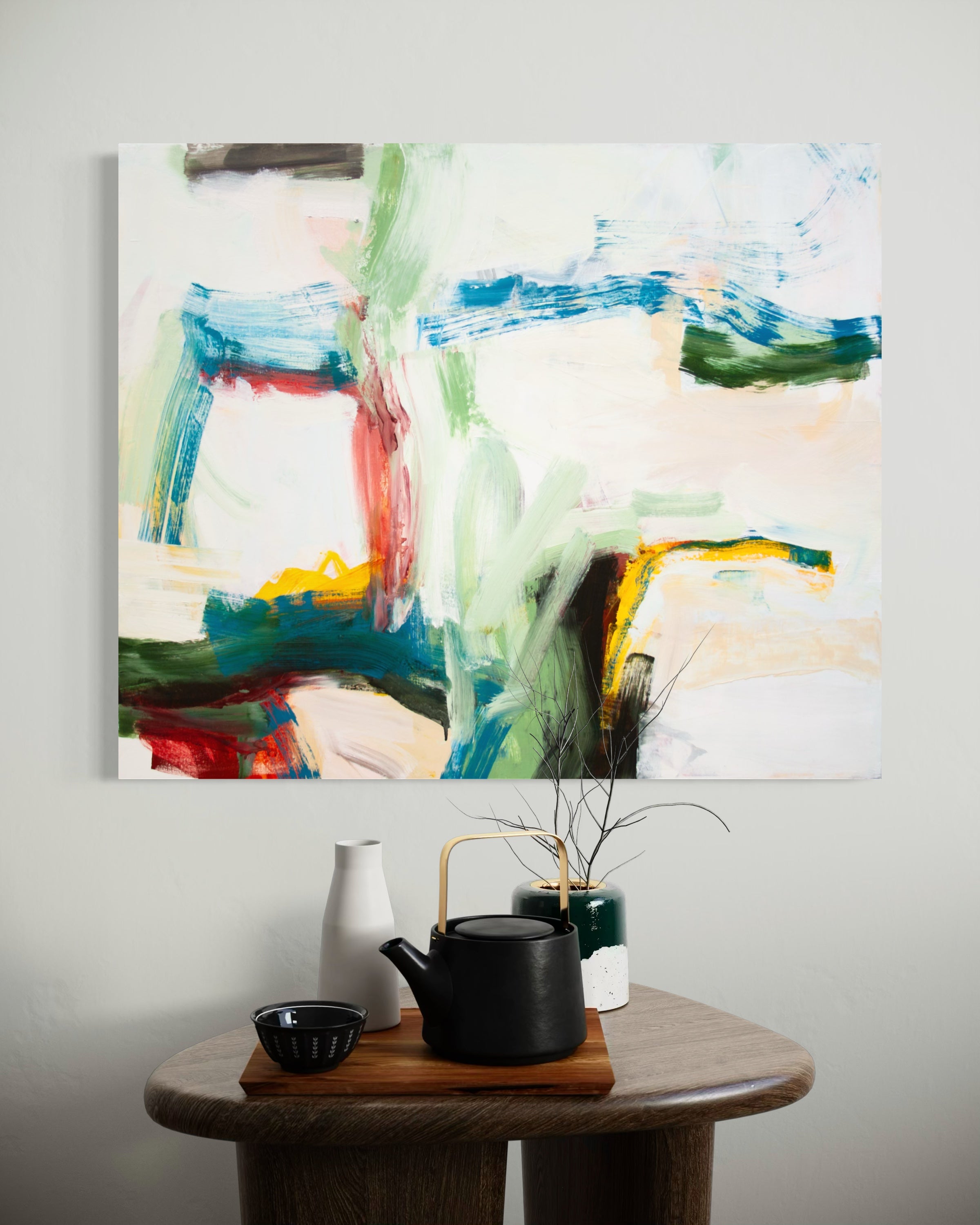 An abstract canvas painting with expressive brushstrokes in green, blue, red, yellow, and black on a white background, displayed on a light gray wall above a wooden side table with a black teapot, white vase, and small bowls.