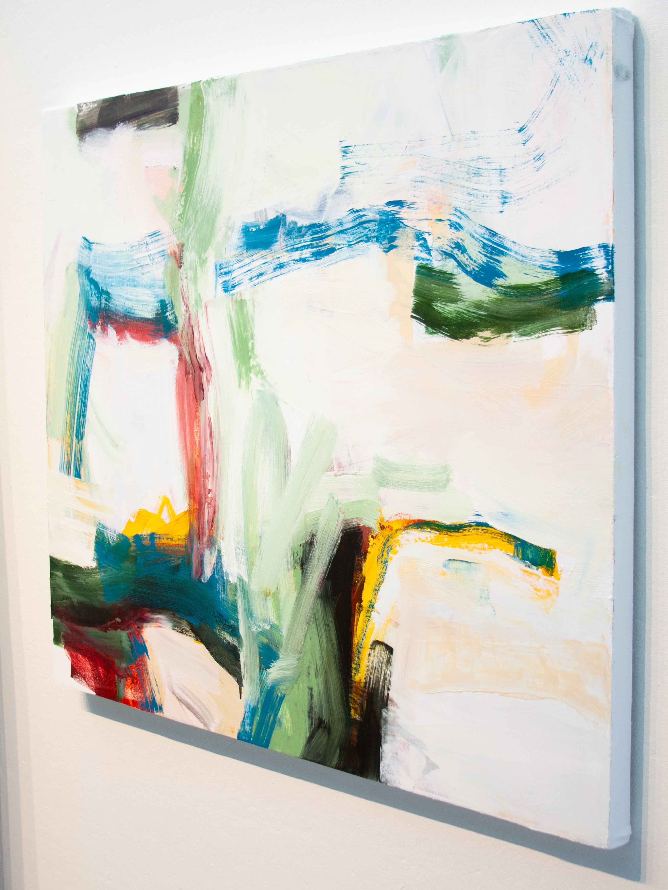 An abstract canvas painting with expressive brushstrokes in blue, green, red, yellow, black, and cream tones, shown hanging on a light gray wall.