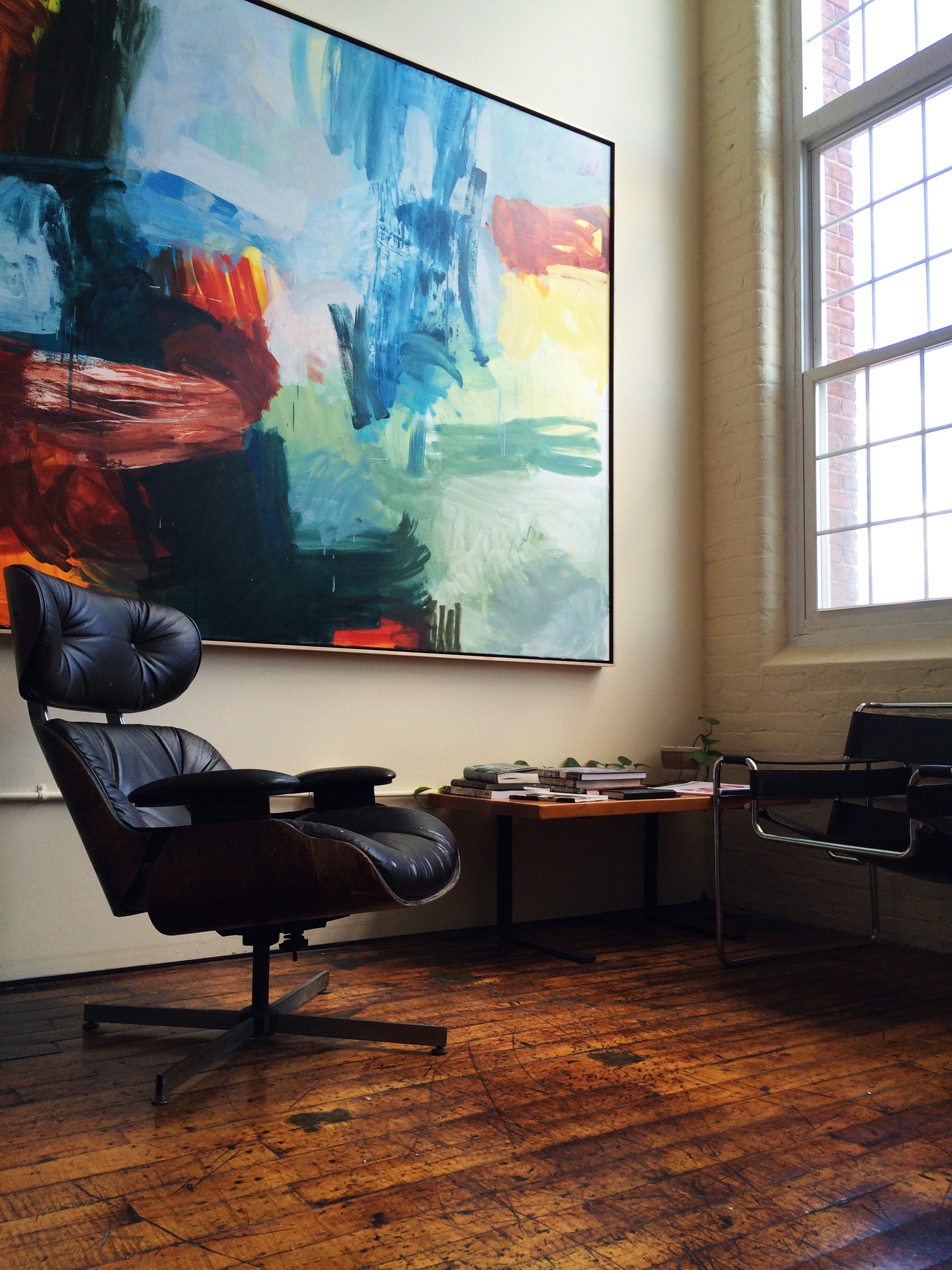 A large abstract canvas painting with blue, green, red, yellow, and black brushstrokes hangs on a beige wall in a modern office setting. In front of the painting is a dark brown leather Eames-style lounge chair, with a small wooden desk and another chair nearby, all set on a rustic hardwood floor.