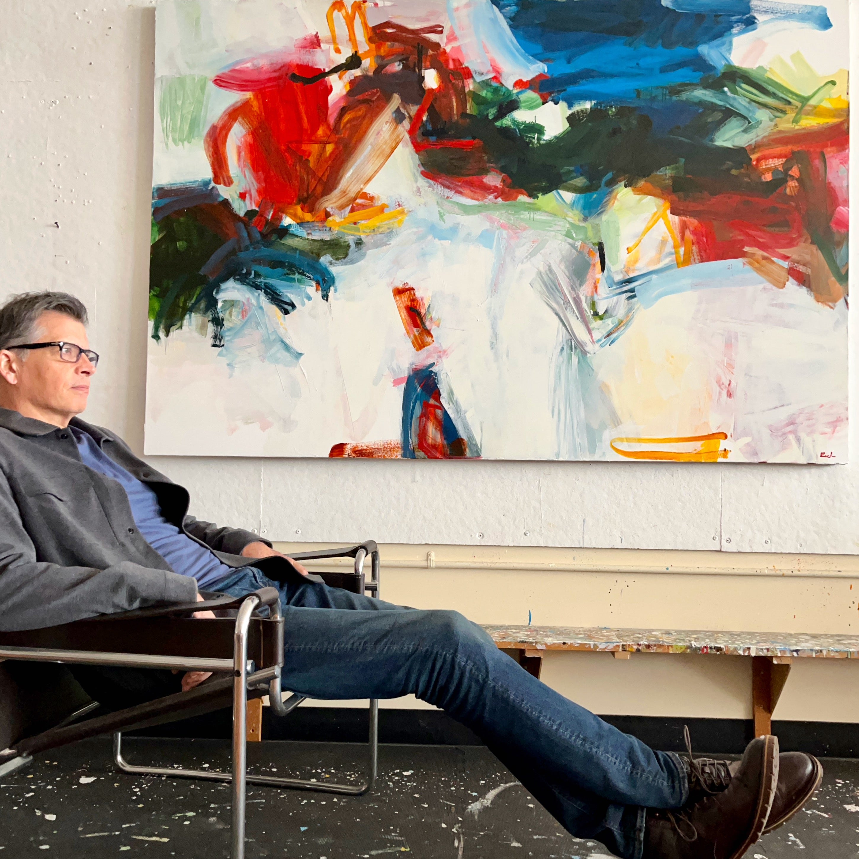 Artist, Michael Rich sits in a modern metal chair in front of a large abstract canvas painting with bold red, blue, green, black, and white brushstrokes.