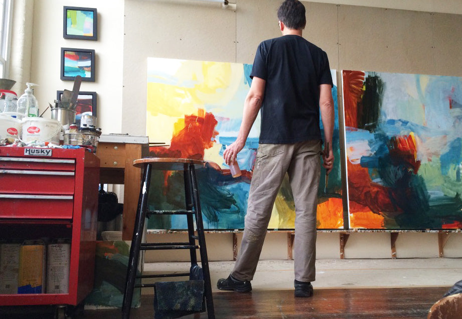 A man stands with his back to the camera in an artist studio, facing two large abstract paintings. He is wearing a black t-shirt and khaki pants.