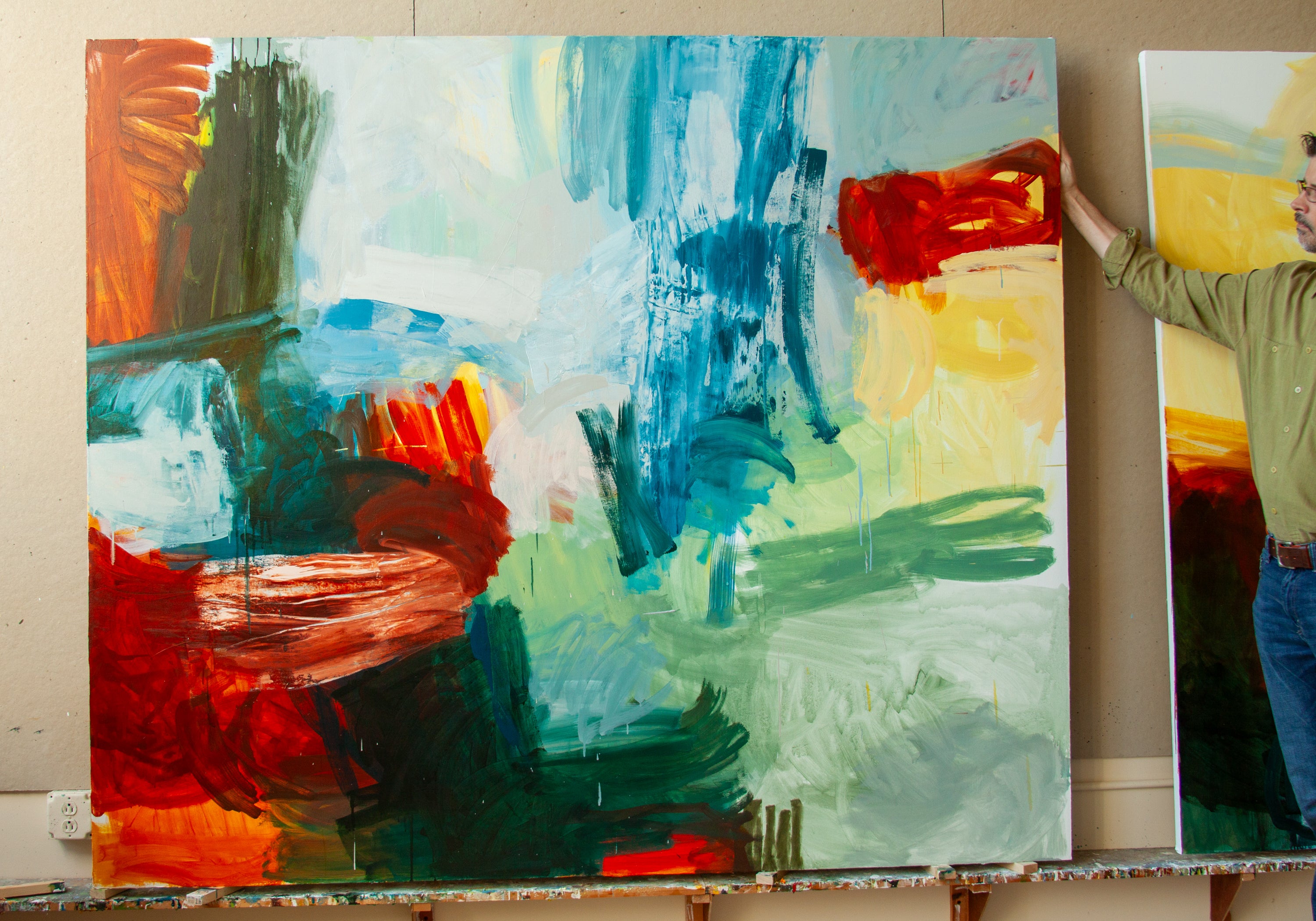 A large abstract canvas painting with bold brushstrokes in blue, green, red, orange, and white, shown indoors with part of a person standing beside it to hold the canvas.