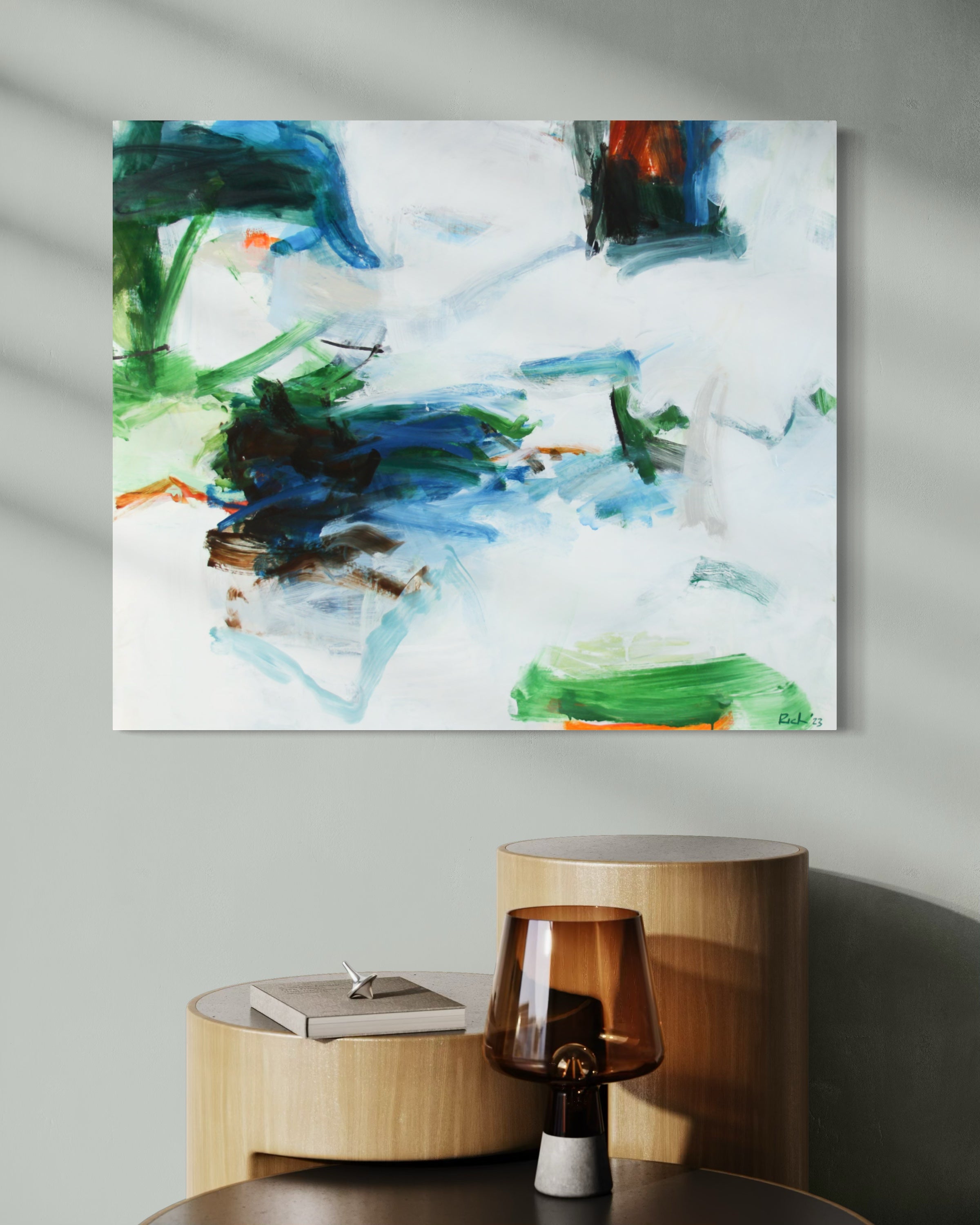 An abstract canvas painting with blue, green, black, and white brushstrokes, displayed on a light gray wall above a wooden side table with a glass lamp.