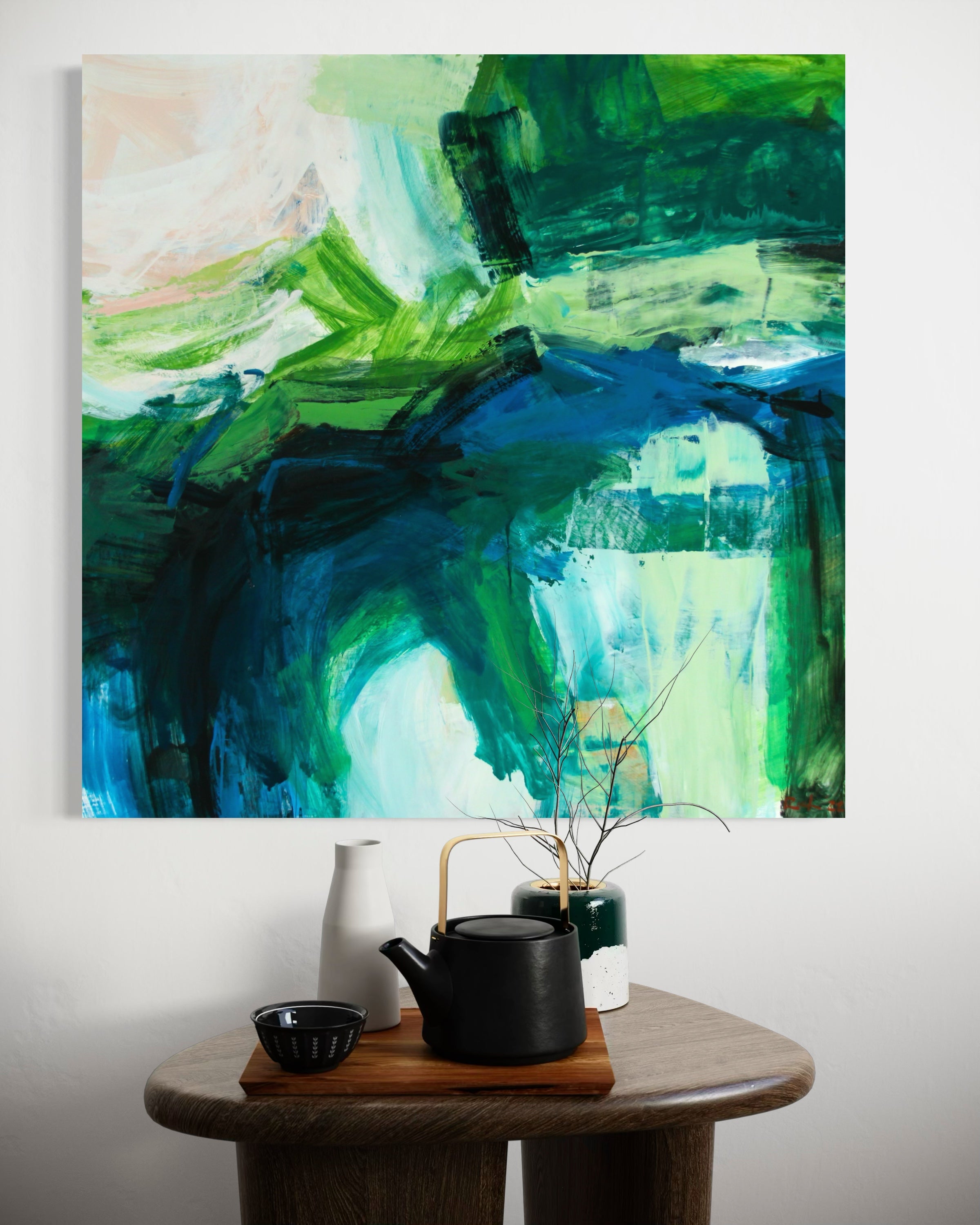 An abstract canvas painting with expressive brushstrokes in shades of green, blue, and white, hanging on a light gray wall above a wooden side table styled with a black teapot, a white vase, and a small bowl.