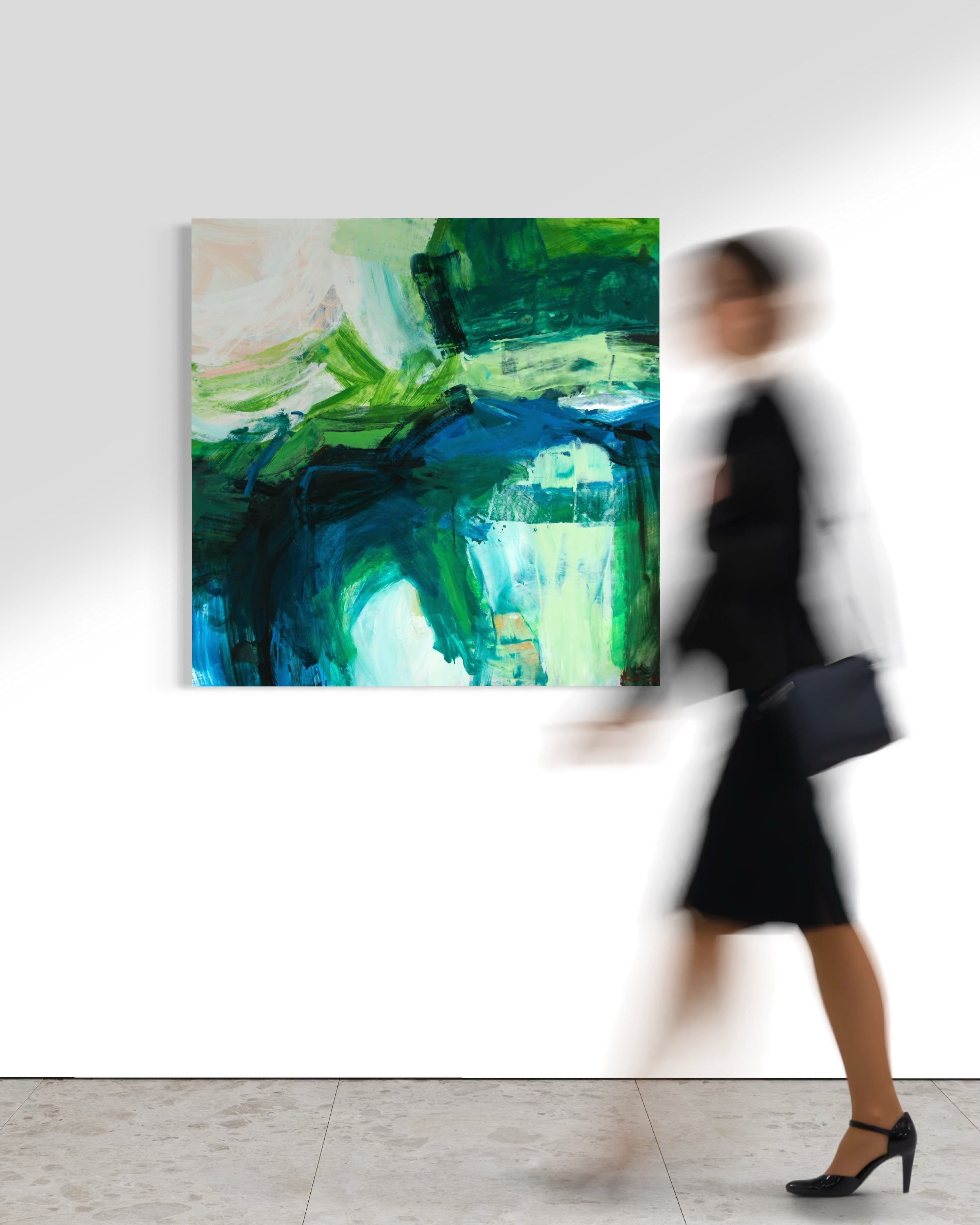 An abstract canvas painting with vibrant green, blue, and white brushstrokes hanging on a white gallery wall, with a blurred woman walking past in the foreground.