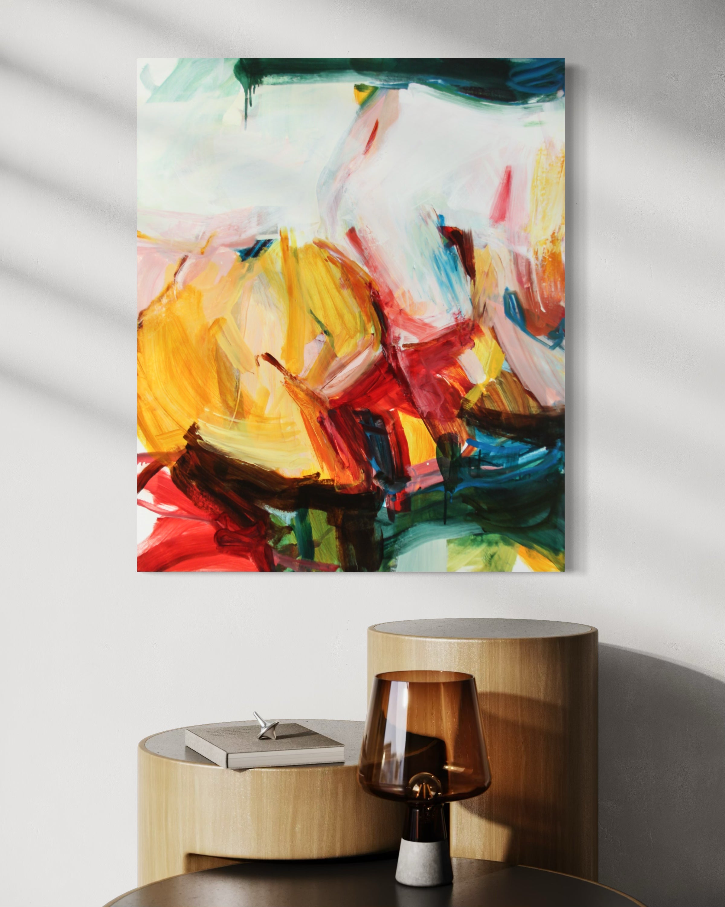 Abstract canvas wall art featuring expressive brushstrokes in yellow, red, blue, green, and white, displayed on a light gray wall above a wooden side table with a lamp and book.