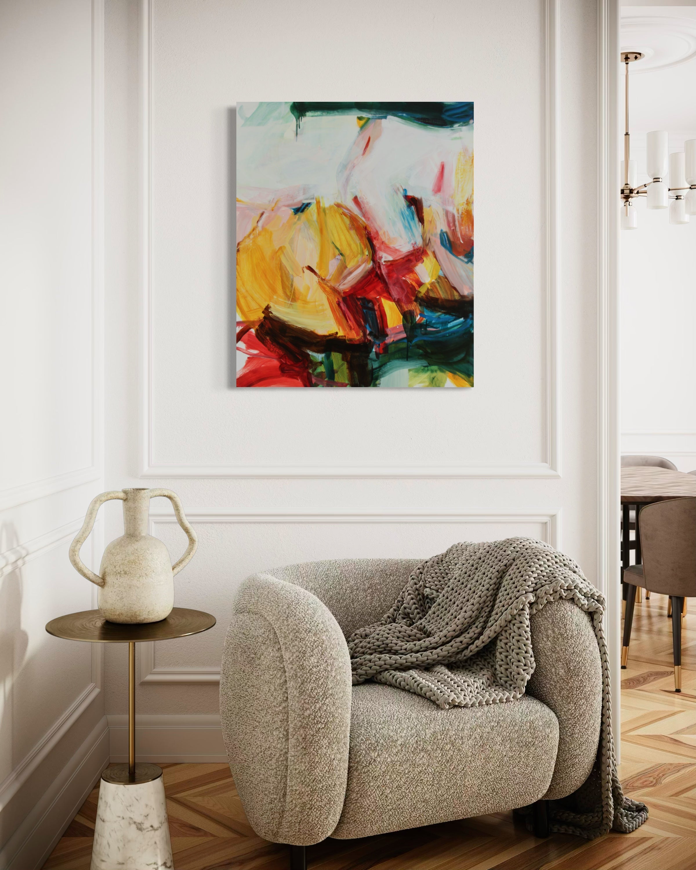 An abstract canvas painting with vibrant yellow, red, blue, and green brushstrokes hanging on a white paneled wall above a beige armchair with a gray knit throw blanket.