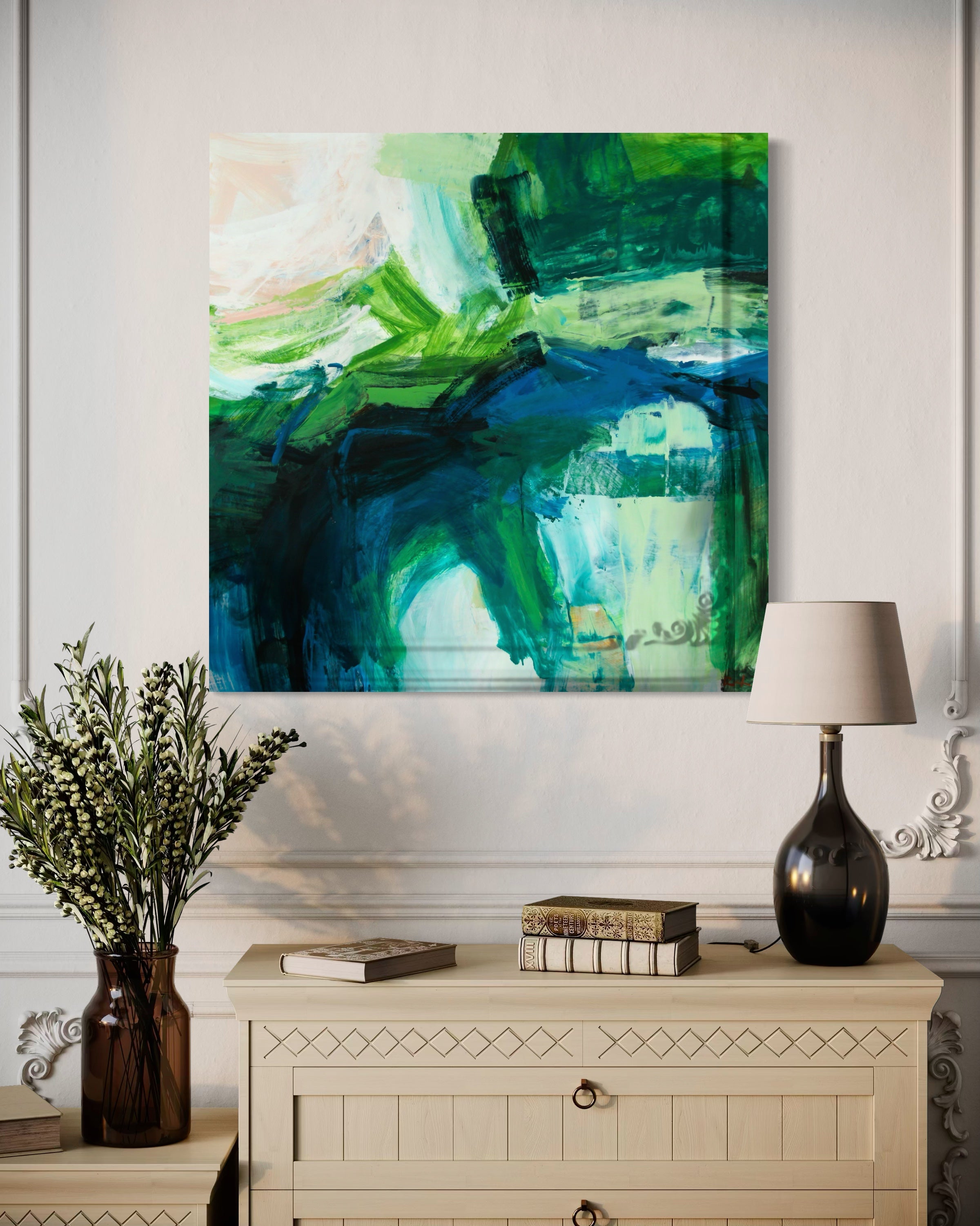 An abstract canvas painting featuring vibrant green, teal, and blue brushstrokes, displayed on a white wall above a cream-colored dresser with a lamp and a vase of greenery.