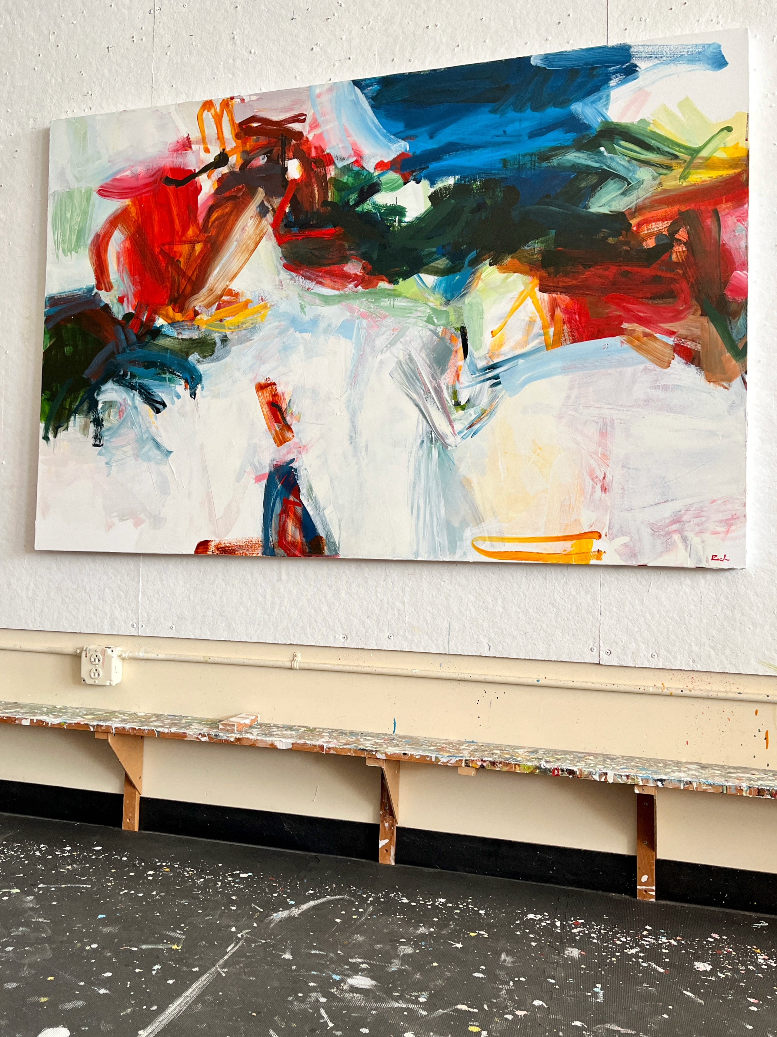 An abstract canvas painting with bold brushstrokes in blue, red, green, yellow, black, and white, displayed on a textured white wall above a workbench.