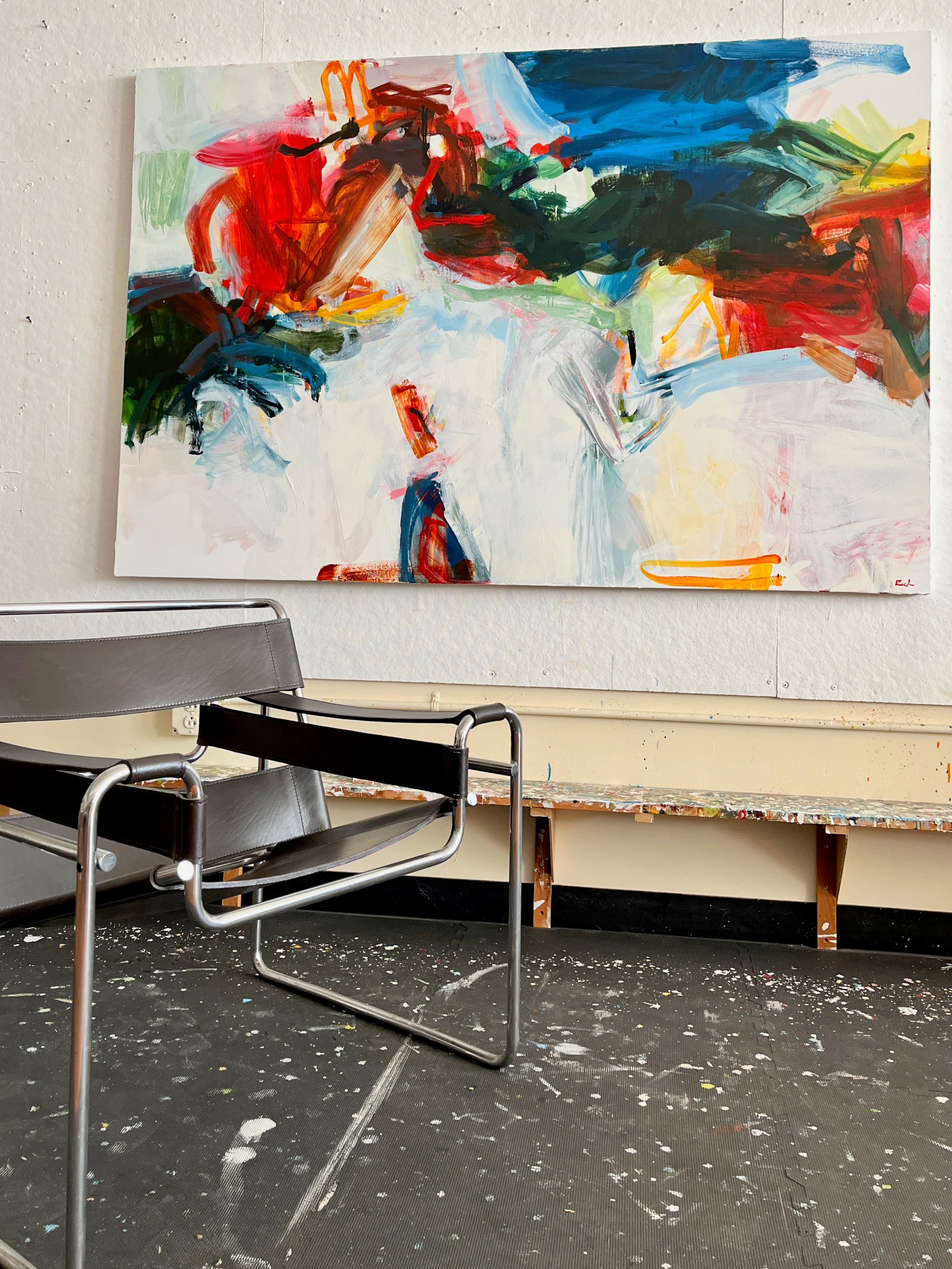 An abstract canvas painting with bold brushstrokes of red, blue, green, yellow, and white hangs on a textured white wall above a modern metal chair in a studio-like interior.
