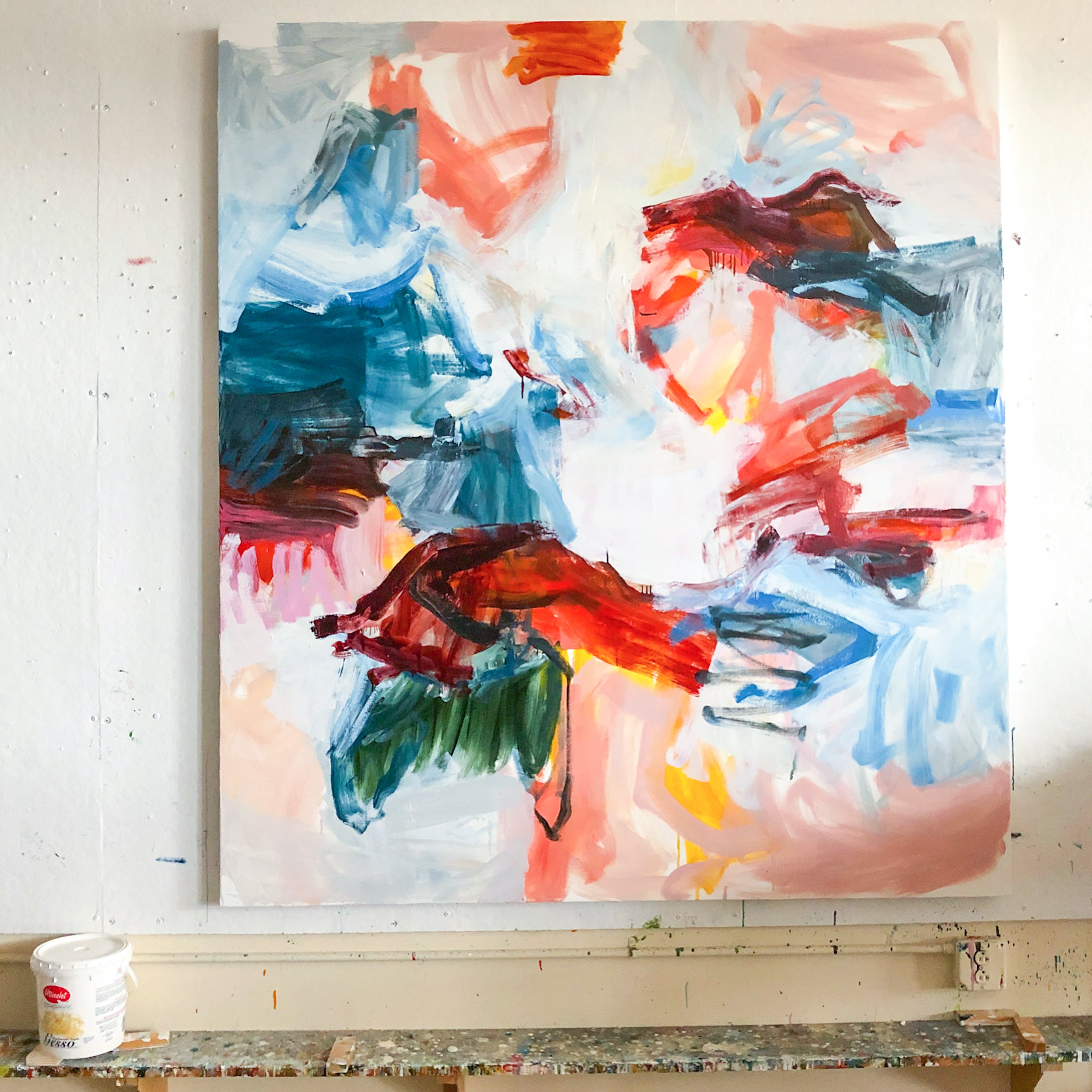 An abstract canvas painting with expressive brushstrokes in blue, red, pink, green, and white, hanging on a white wall above a paint-splattered workbench. A white paint bucket is visible on the lower left.