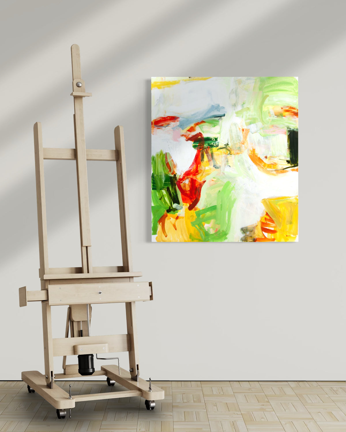 An abstract canvas painting by Michael Rich with vibrant green, yellow, red, and white brushstrokes hanging on a light gray wall next to a wooden easel.